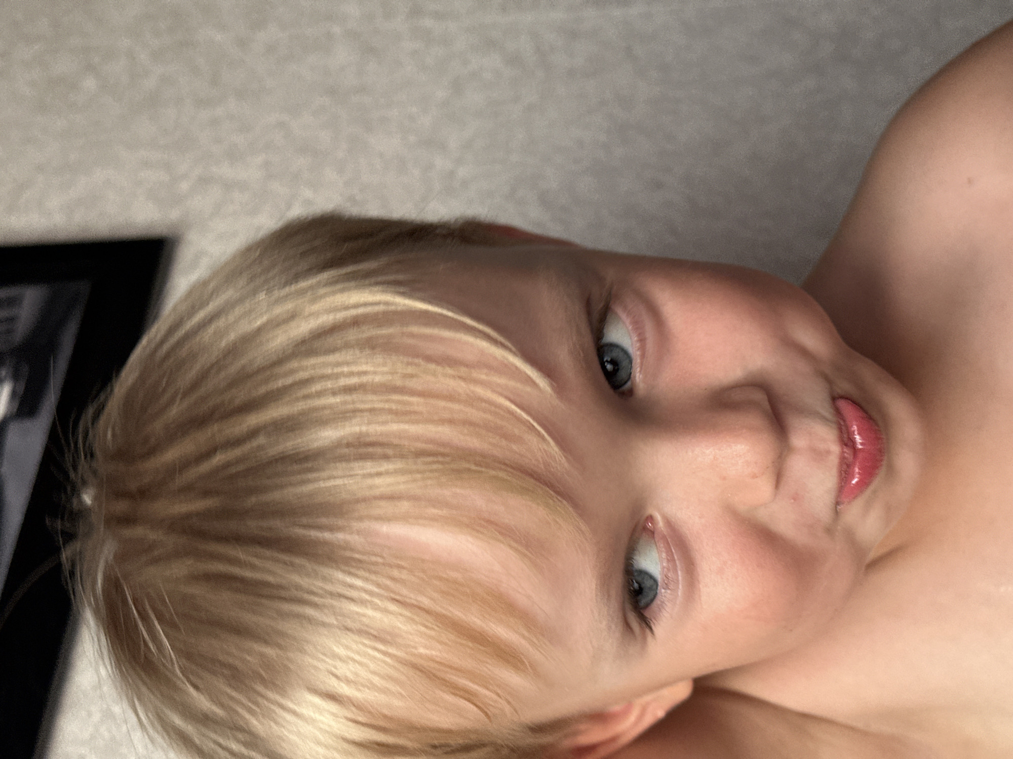 A close-up, low-angle, portrait-style photo shows a young child with blonde hair. The child is lying down and looking towards the camera. Their eyes are blue and open, with one eye more visible than the other. The child's mouth is slightly open with their tongue peeking out, giving them a playful expression. The lighting suggests it might be daytime. The background appears to be a textured wall, possibly in a home setting in Energetik, Russia. There are no visible objects other than the child and the background. The overall mood is lighthearted and candid.