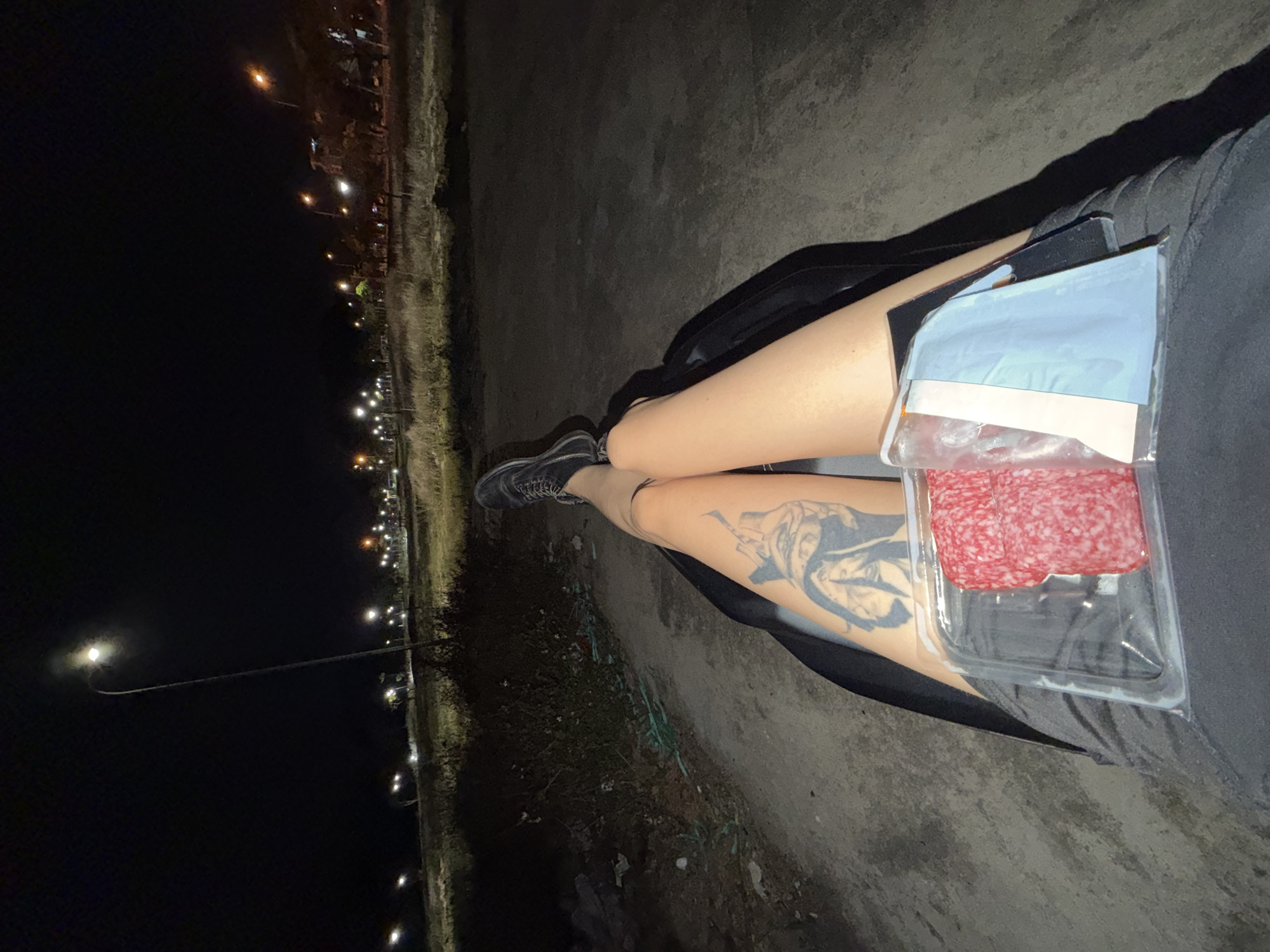 A ground-level, eye-level photograph taken at night shows a person sitting with their legs extended. The person is wearing black shoes and dark-colored clothing. A package of sliced meat rests on the person's legs. A tattoo is visible on the upper part of one leg. The background consists of a paved area and sparse vegetation. Streetlights illuminate the area. The location cannot be determined to be in Đà Nẵng, Vietnam, solely based on the visual information in this image.
