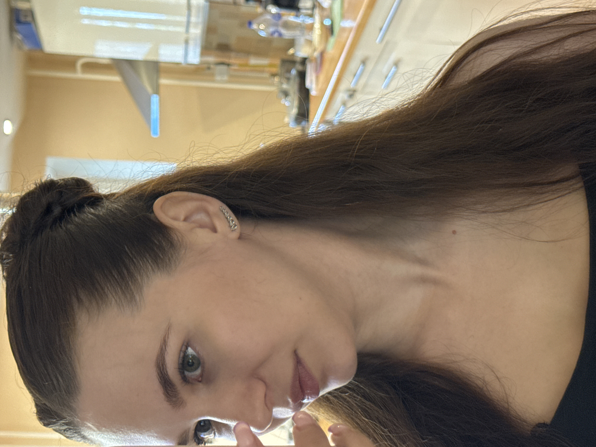 A close-up portrait shows a young woman indoors. The image is rotated 90 degrees clockwise, presenting her head tilted significantly to the right. She has long, dark brown hair, part of which is gathered in a bun on the upper left side of her head, with loose strands cascading over her shoulder and chest. Her green-hazel eyes gaze directly at the camera, complemented by well-defined eyebrows. Her lips are slightly parted and appear glossy.
She wears a silver-toned earring, possibly a small hoop or cuff with decorative stones, on her visible right ear. Her skin is clear, and a small mole is visible on her neck/shoulder area. A dark, likely black, garment covers her shoulder and chest. The tips of her fingers are partially visible near her lower face.
The background is an indistinct, blurry indoor setting with light beige or pale yellow walls. Reflections of overhead lighting or windows are visible, along with vague shapes that suggest shelves or counters holding various items, possibly bottles or containers. Some elements in the background appear to be light sources or signs with a blue hue. The overall impression is a casual, indoor environment, potentially a commercial space. There is no visible text in the image. The location context provided is Chaikovsky, Russia.