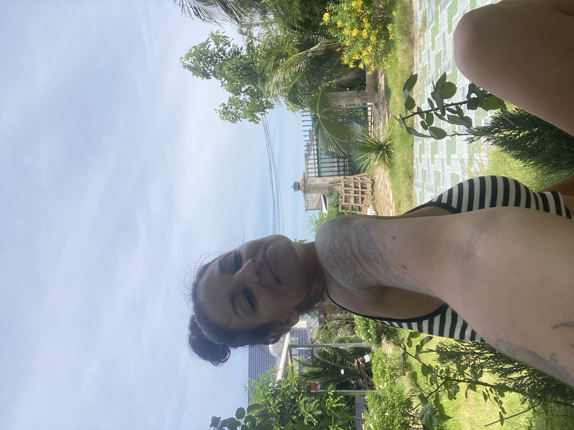 A woman with dark hair pulled into a bun is taking a selfie in a garden. She is wearing a black and white striped top and has tattoos on her arm. The background shows lush greenery, a tiled patio, a fence, and a building with a blue roof. The sky is overcast. The scene appears to be in a residential area of Mũi Né, Vietnam.