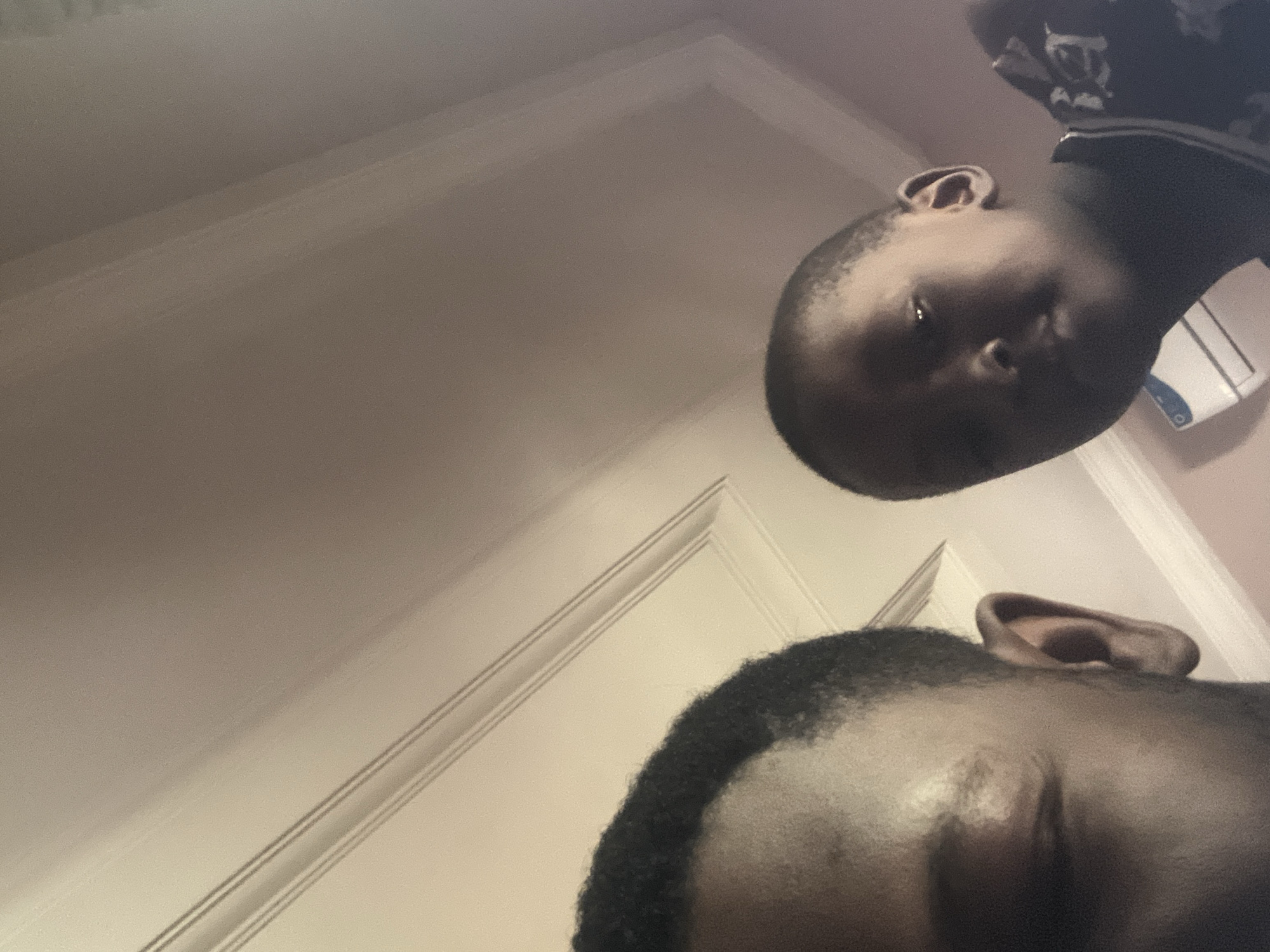 The image shows a close-up, low-angle shot of two individuals, an adult and a child, looking towards the camera. The adult, on the left, has dark hair and is partially visible, with only the side of their face, including their eye and ear, in frame. The child, on the right, is in focus and appears to be a young boy with short hair. He is looking directly at the camera with a neutral expression. Both individuals have dark skin tones.
The background consists of a wall with ornate molding or trim, suggesting an indoor setting, possibly a home or a building. The lighting appears to be overhead, casting shadows and highlighting the contours of their faces. There is a light-colored object, possibly a vent or a piece of electronic equipment, visible in the lower right portion of the frame.
Given the limited visual information, it is impossible to determine the specific location within Jalingo, Nigeria. The image captures a candid moment, focusing on the faces of the two subjects and their immediate surroundings.