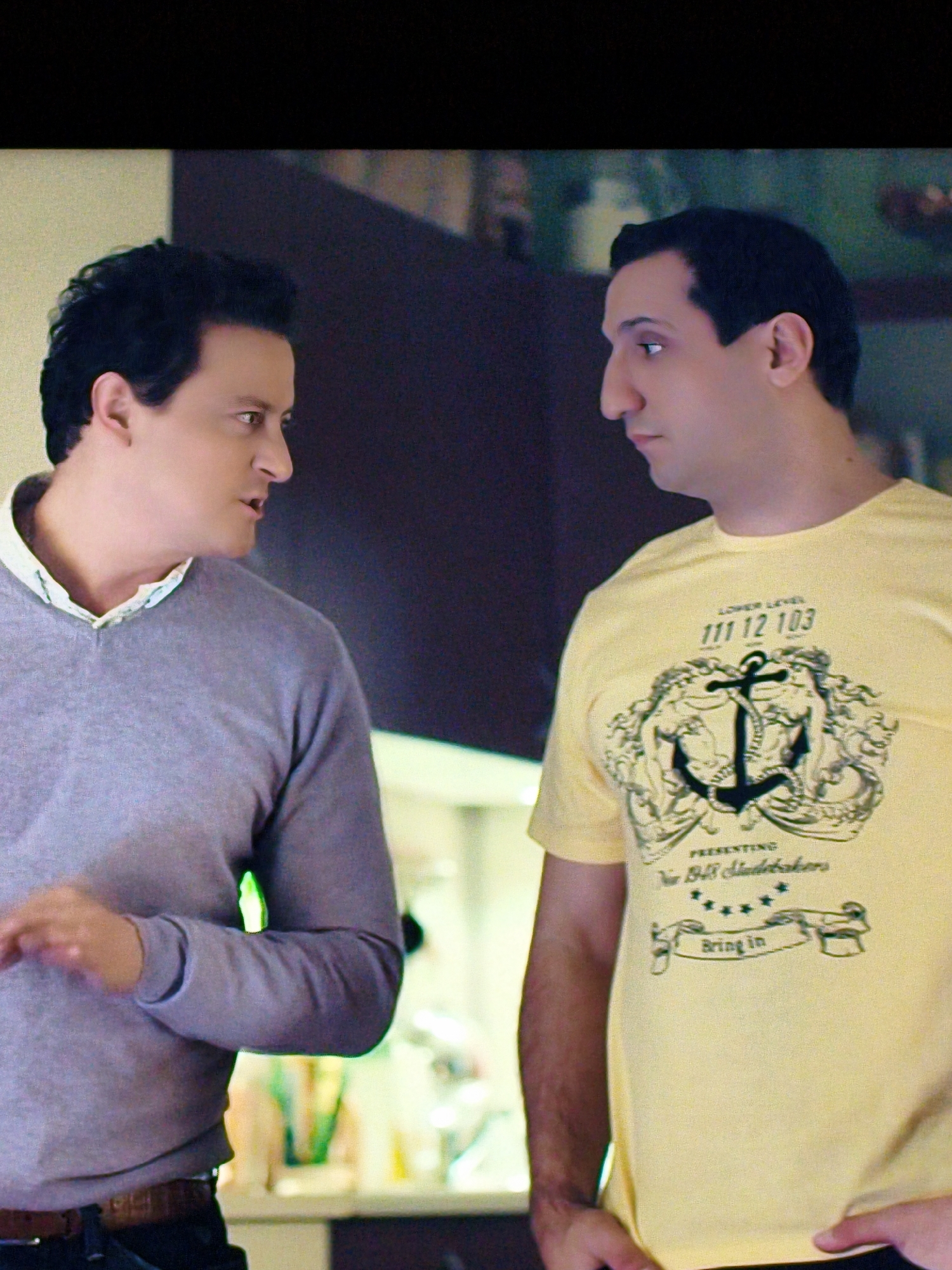 Two men stand facing each other indoors. The man on the left, with dark curly hair and light skin, wears a grey v-neck sweater over a white collared shirt. He is gesturing with his right hand as he speaks. The man on the right, with dark straight hair and light skin, wears a yellow t-shirt with a graphic design featuring an anchor, two figures, stars, and text that reads "LOWER LEVEL 111 12 103", "PRESENTING New Bell Studebakers", and "Bring In". The background shows a dark wall and a blurred interior space with some light sources. This scene is not set in Vladivostok, Russia.
