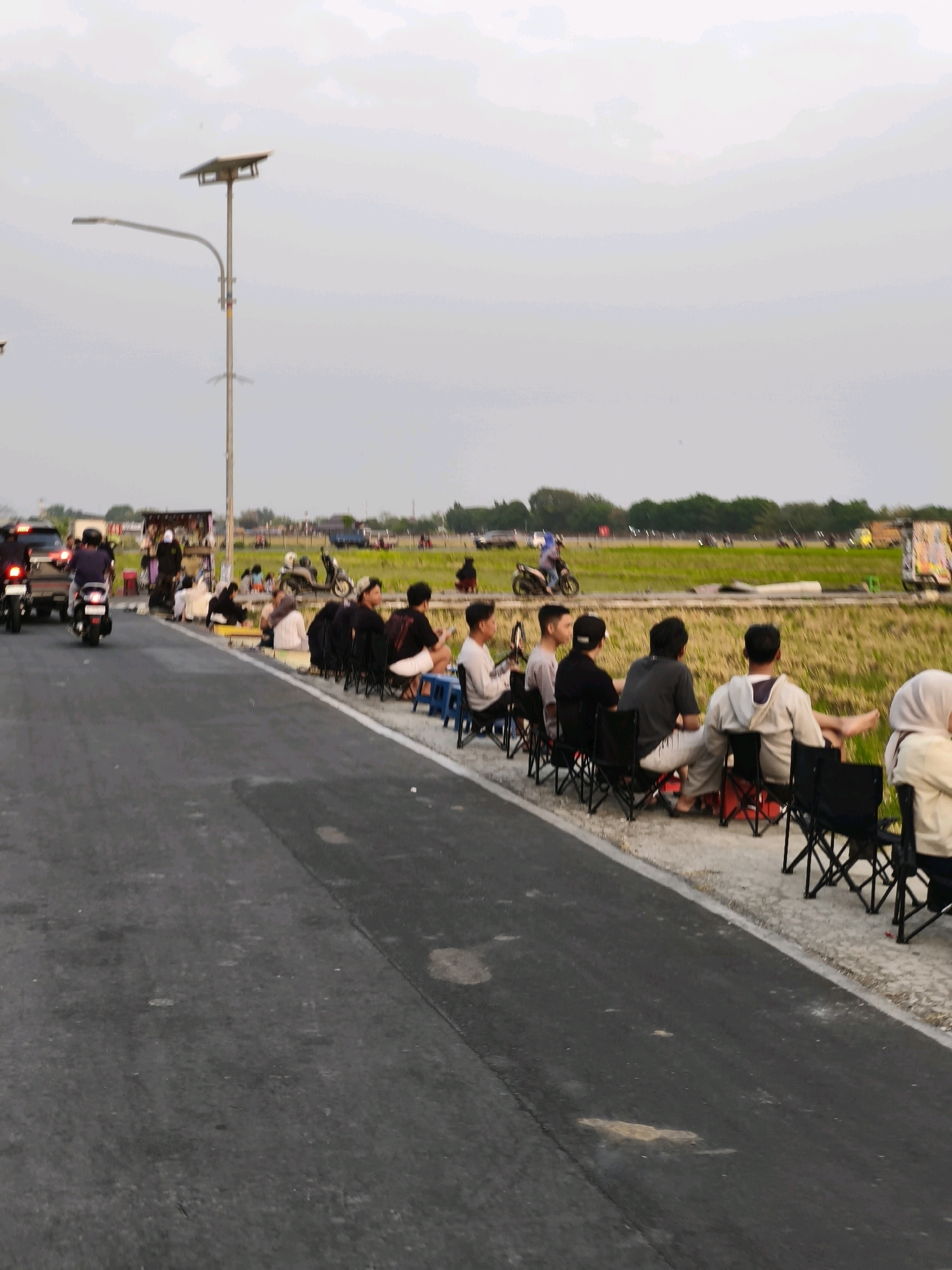 The image depicts a street scene in Desa Ngesrep, Indonesia, likely during the late afternoon or early evening, given the lighting conditions. The foreground features a paved road. Adjacent to the road, on the right side of the image, a group of people are seated on folding chairs, facing a field of dry, yellowing crops. The individuals are casually dressed, and their attention appears to be directed towards something in the distance.
Further down the road, on the left side, there's a collection of roadside stalls or carts, with more people gathered around them. Several motorcycles are visible, some being ridden by individuals wearing helmets, while others are parked. A street lamp with a solar panel is prominent in the background. Beyond the immediate roadside area, a vast expanse of agricultural land stretches out, with trees forming a distant treeline. The sky is overcast with a pale, diffused light. The overall impression is one of a community gathering or a local event taking place alongside a road bordering a rural landscape.