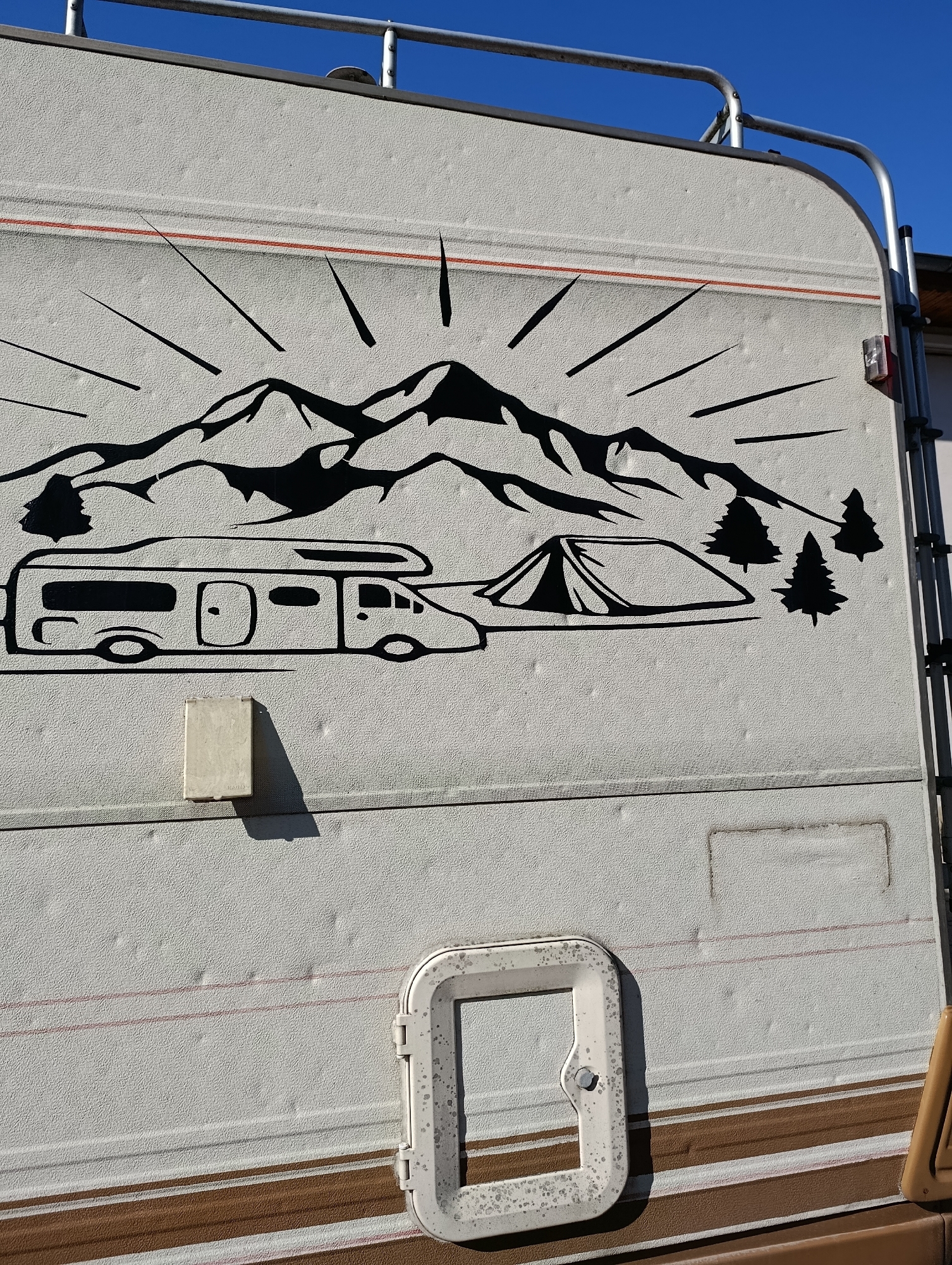 The image shows a side view of a white RV with a black decal depicting a mountain range, a camper van, a tent, and trees. The decal is applied to the textured surface of the RV's exterior. A metal railing runs along the top edge of the RV. A red stripe runs horizontally across the RV's side above the decal. Below the decal, there is a cream-colored rectangular vent cover with a shadow cast on the RV. Further down, there is a brown stripe and a white window frame with a latch. The RV appears to be parked outdoors under a clear blue sky. The location is identified as Razgrad, Bulgaria.