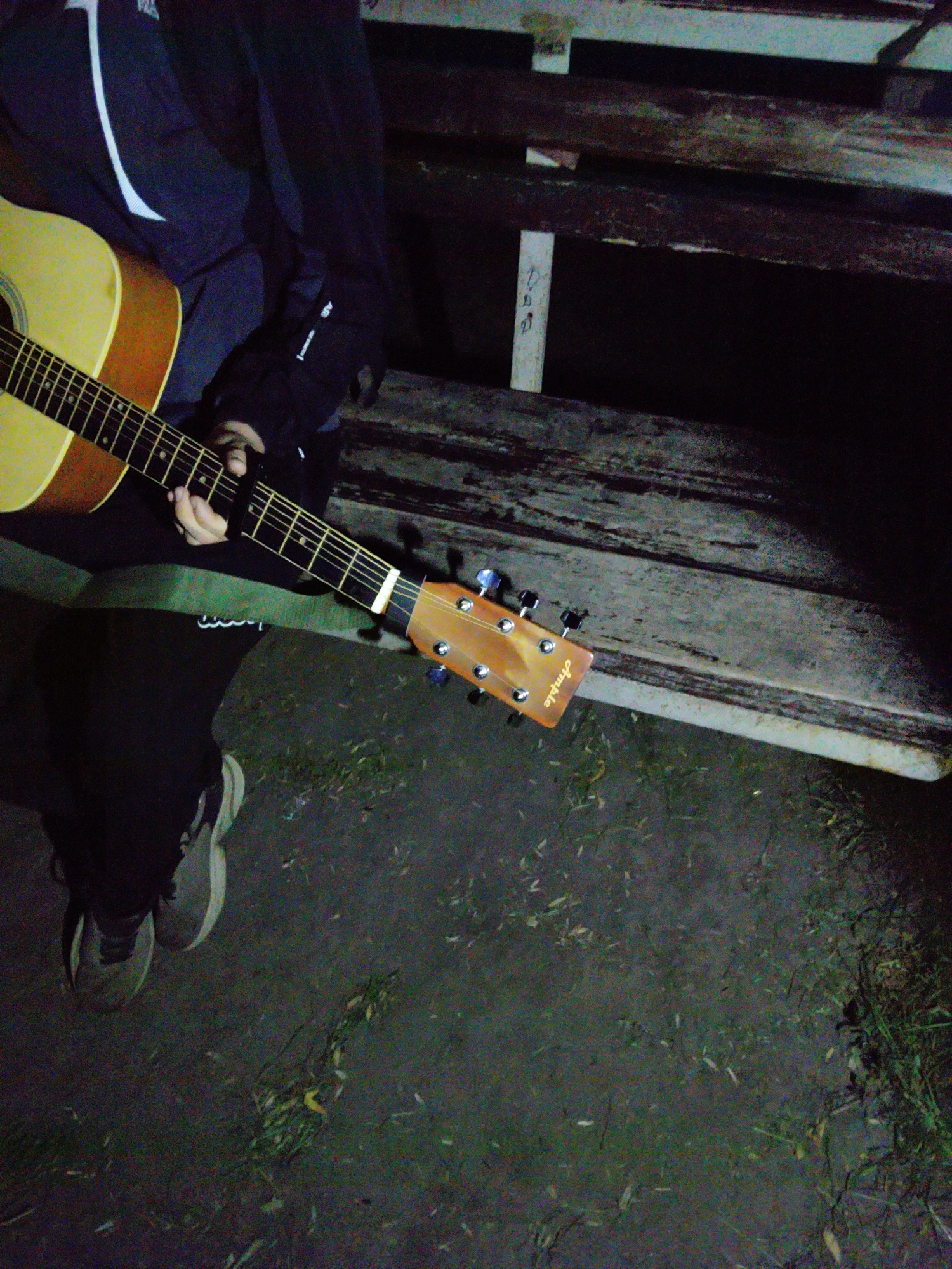 An overhead, dimly lit image shows a person playing an acoustic guitar outdoors at night in Ustyvytsia, Ukraine. The individual, dressed in a dark jacket with a white stripe, black pants, and grey and white athletic shoes, is seated, likely on a wooden bench which is partially visible behind and to their right.
The light-colored acoustic guitar, with a dark fretboard, is held across the person's lap. A black capo is clamped onto the guitar's neck, indicating preparation for playing in a specific key. The person's left hand is positioned on the fretboard, suggesting they are forming chords. The guitar's headstock, featuring chrome tuning pegs, is visible on the right side of the frame, with the brand name "Ample" clearly inscribed vertically.
The setting appears to be an outdoor area, possibly a park or garden, with a rustic wooden bench providing seating. The ground beneath the person is dark and uneven, with scattered debris and what appears to be grass or dirt. The overall low light levels suggest it is nighttime, creating a quiet and intimate atmosphere. No specific emotions are discernible from the partial view of the person.