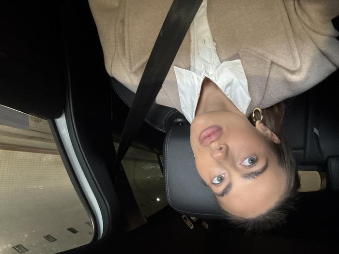 A female individual is depicted in a close-up selfie taken from the passenger seat of a modern vehicle. She has dark hair pulled back, light-colored eyes, prominent eyebrows, and full lips, with gold hoop earrings. She is wearing a white collared shirt beneath a beige overcoat and is secured by a black seatbelt diagonally across her chest. The car's interior features black upholstery with visible white stitching on the seat and headrest. Through the vehicle's window, a well-lit road tunnel is visible in the background. The tunnel walls are light-colored, appearing to be tiled or paneled with a horizontal pattern, and illuminated by rectangular overhead light fixtures. This tunnel is identified as the Wisłostrada Tunnel (Tunel pod Wisłostradą), specifically the section beneath Plac Zamkowy, located in the city of Warsaw, Poland.