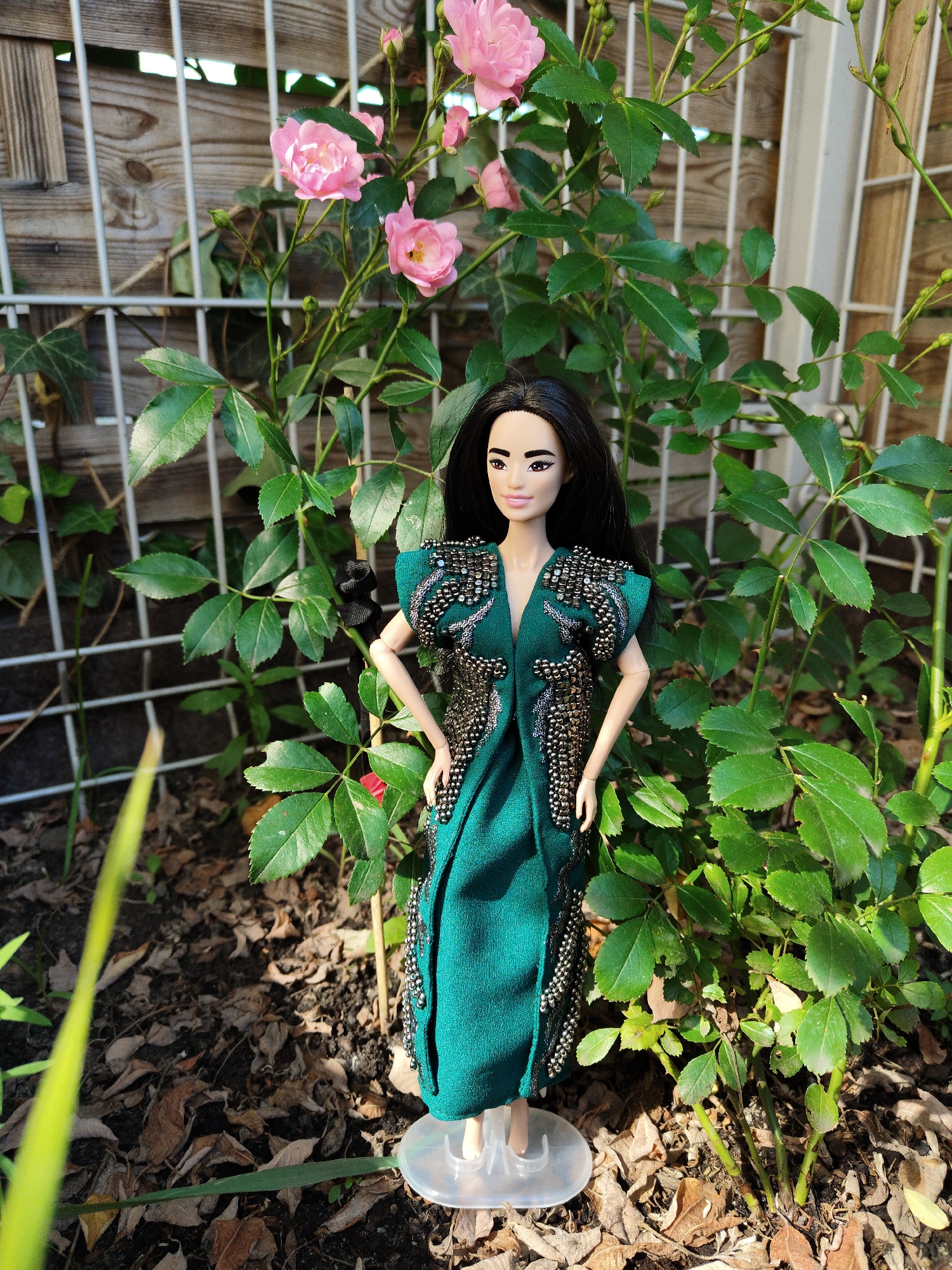 A doll, dressed in an ornate teal gown embellished with silver beads, stands in a garden setting, partially obscured by green foliage and pink roses. The doll has long black hair and is positioned on a clear plastic stand. Behind the doll, a white metal fence with a wooden backing is visible. The ground is covered with dry leaves and soil. The scene is illuminated by sunlight, casting shadows. The image was taken in Wrocław, Poland.