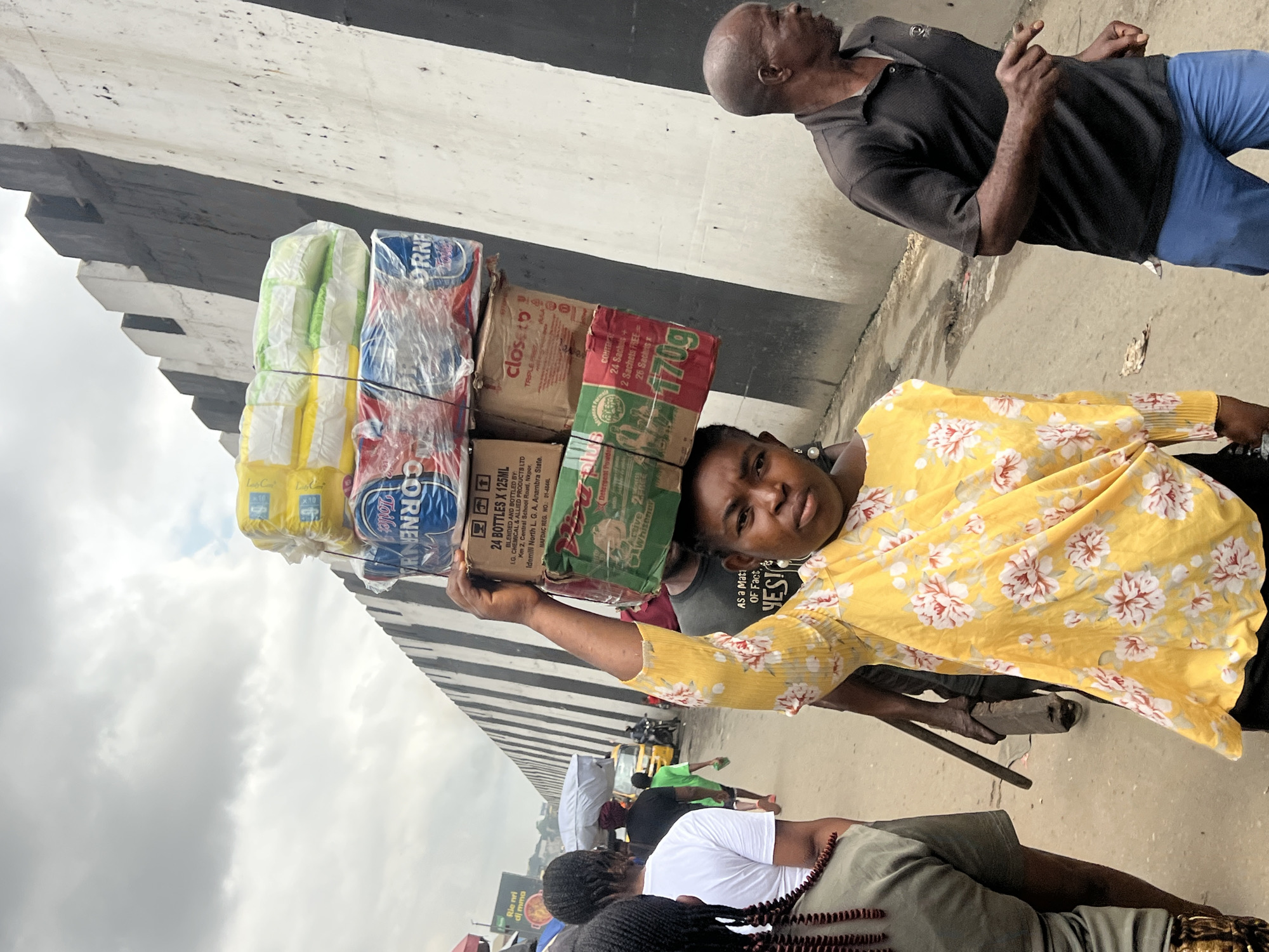 The image depicts a street scene in Nkpor, Nigeria, under a cloudy sky. A woman in a yellow floral print top balances a stack of goods on her head, including packaged diapers and detergent. She is walking towards the viewer, her face partially obscured by the merchandise. To her right, a man in a dark polo shirt and blue pants is walking away, gesturing with his right hand. In the background, other people are visible, some carrying items on their heads, and a yellow car is parked on the street. A tall, black and white striped wall runs along the right side of the image.