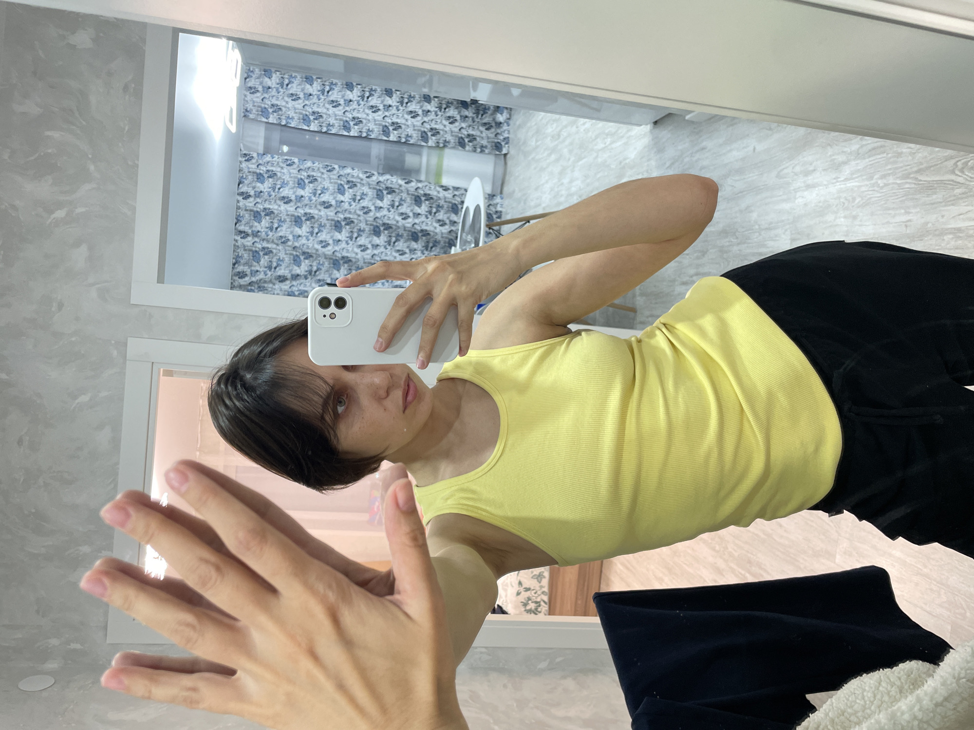 A person is taking a mirror selfie. The person has dark hair and is wearing a yellow tank top and black pants. The background shows a room with patterned curtains, an ironing board, and a marble-like wall. The location is possibly in Izhevsk, Russia.
