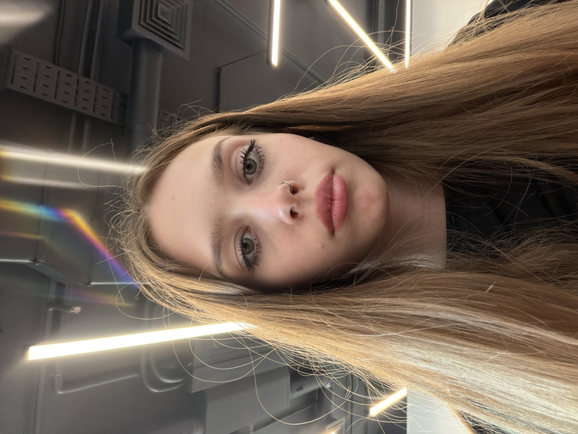 This is a close-up, angled selfie of a young person with long, light brown hair. The subject has green eyes, full lips with a pink tint, and a small nose ring. Their eyebrows are well-defined, and their eyelashes appear to be enhanced with mascara. The background is dark and industrial, featuring what appears to be a metal ceiling or wall with vent-like structures and bright, linear light fixtures. A prism-like light reflection in rainbow hues is visible on the left side of the image. The lighting suggests an indoor environment, possibly an office or a modern building. The subject's expression is neutral, and the overall mood is casual, consistent with a selfie. There is no visible text in the image.