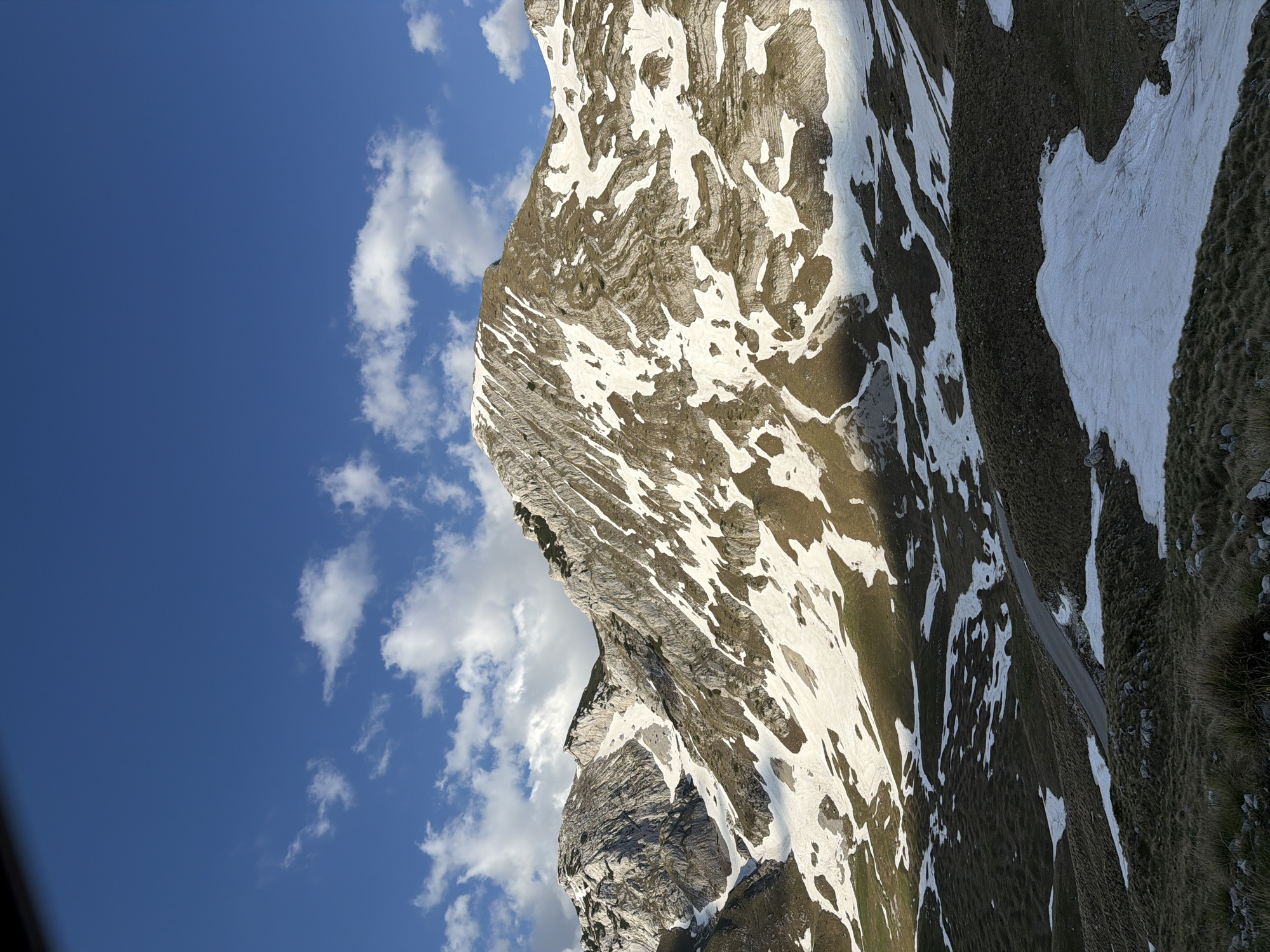 A landscape in Боричје, Montenegro shows a mountain range with a mix of exposed rock, snow patches, and grassy areas under a partly cloudy blue sky. A road can be seen snaking up the side of the mountain.