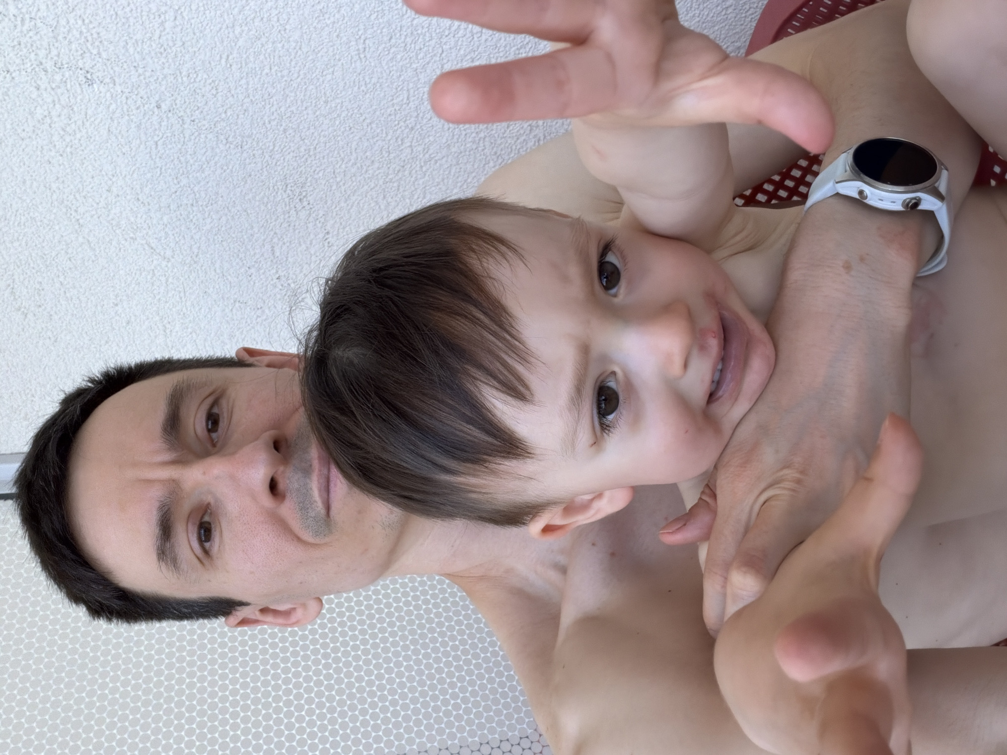 A man and a child, possibly his son, are posing for a selfie. The man is holding the child close, and both are looking at the camera. The man is shirtless and wearing a smartwatch. The child is also shirtless. The background appears to be a white wall and a red patterned surface. There is no information in the image to determine if the photo was taken in Sochi, Russia.