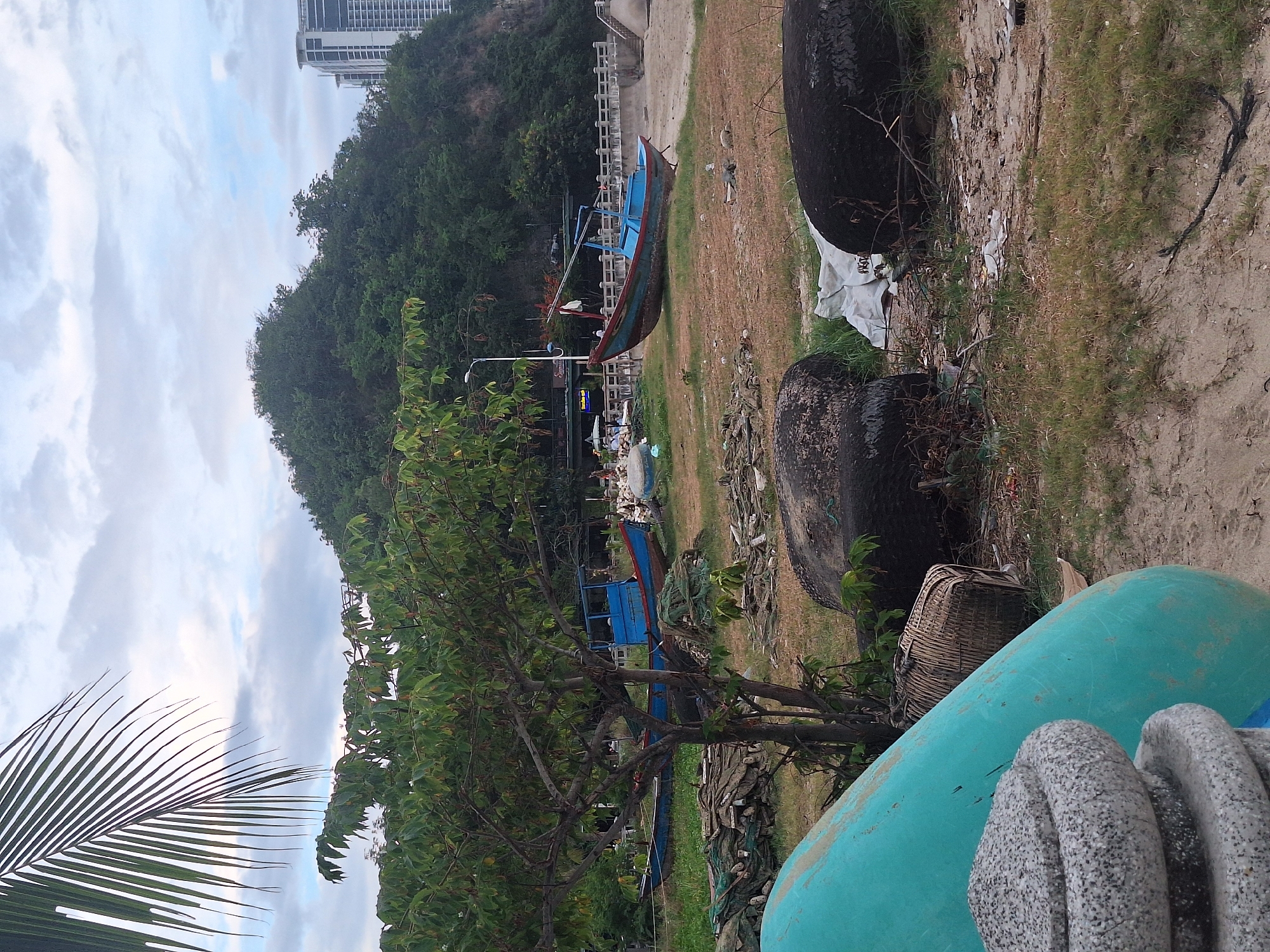 The image depicts a coastal scene in Nha Trang, Vietnam, likely on a beach or foreshore area. In the foreground, a large, teal-colored object, possibly part of a boat or construction material, dominates the left side. Next to it, a stack of grey, circular objects, possibly decorative stone elements or weights, are visible. Behind these, a woven basket lies on the ground.
The middle ground features a grassy area with several large, dark, spherical objects, which appear to be tires. Scattered around them are various debris, including white plastic sheeting, branches, and other refuse. Two blue fishing boats are present; one is closer to the left, partially obscured by a tree, and the other is positioned further to the right. Fishing nets and wood piles are also visible. A small tree stands in the center, providing shade.
In the background, a lush green hill rises, covered in dense vegetation. At the top of the hill, modern buildings with white facades and many windows are visible, indicating an urban or developed area adjacent to the natural landscape. A white railing or bridge structure runs along the base of the hill, suggesting a walkway or embankment. The sky is overcast with scattered clouds, and a palm frond extends from the top left corner of the image, framing the scene. The overall impression is one of a working coastal area, possibly a fishing village or a staging ground for marine activities, with elements of both natural beauty and human activity.