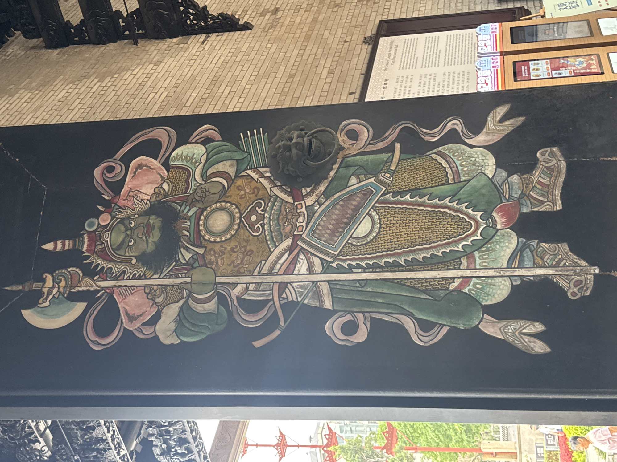 The image depicts a colorful, traditional painting of a warrior figure on a dark background, likely part of architectural decoration, possibly a lintel or beam. The warrior is elaborately dressed in armor, carrying a polearm with an axe-like blade. The details of the painting include ribbons or sashes surrounding the figure and ornate designs on the armor. A lion-shaped door knocker is positioned above the warrior.
The painting is located in Liwan District, China. Elements of the surrounding environment are partially visible, including some signage and architectural details. The scene has been photographed from an angle, suggesting the observer is positioned below and looking upwards towards the painted surface.