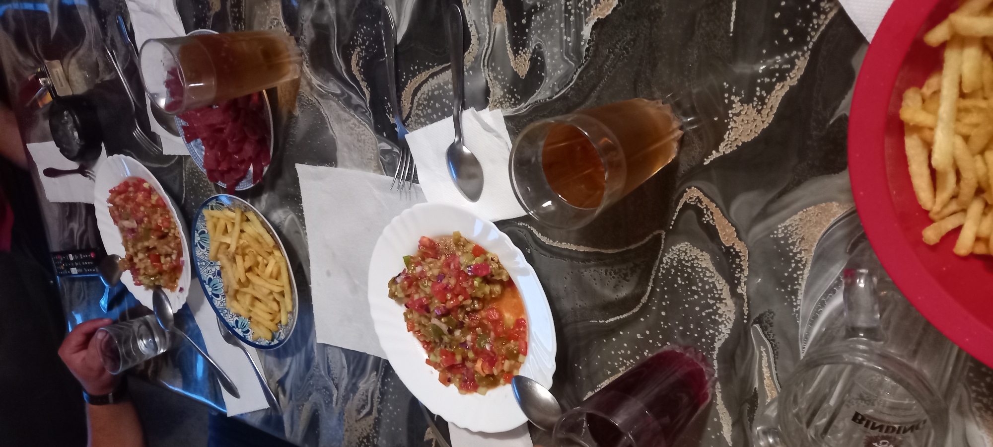 The image depicts a table setting with food and drinks, likely in a restaurant or home. There are several plates of food, including a salad-like dish with chopped tomatoes and other vegetables, a plate of french fries, and another plate with red, thinly sliced food, possibly cured meat or dried fruit. Two glasses of amber-colored liquid are visible, one taller than the other. A glass with a dark red liquid is also present. A person's hand, wearing a watch, is visible on the left side of the frame, reaching for a glass. Utensils such as spoons and forks are scattered on the table, along with napkins. The table surface has a dark, patterned design. A remote control is also visible on the table. The scene appears to be set indoors, with artificial lighting. The location in Iskra, Bulgaria is not discernible from the visual information provided in the image.