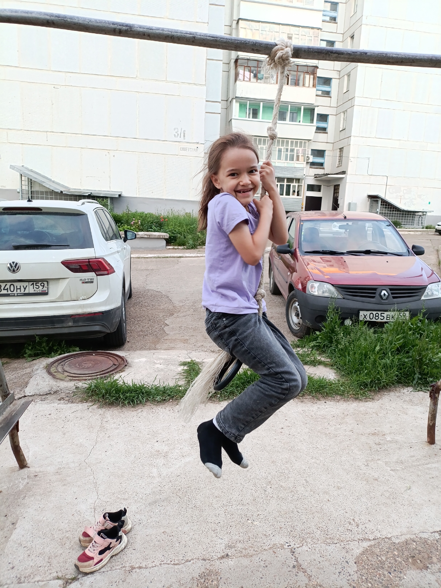 A young girl is captured mid-air on a rope swing in a residential area of Chaikovsky, Perm Krai, Russia. She is centrally positioned, smiling broadly with a joyful expression, and looking directly at the viewer. Her dark hair is swept back, and she wears a light purple short-sleeved t-shirt, dark grey jeans, and black socks with white toes. Both hands firmly grip a thick, natural-fiber rope, which is knotted around a horizontal, dark metal bar positioned above her head. Her feet are off the ground, consistent with the act of swinging.
In the immediate foreground to the lower left, a pair of pink and maroon athletic shoes rest on the textured concrete ground. The ground throughout the scene is primarily cracked concrete, with patches of green grass and dirt, particularly around a circular metal manhole cover to the left and near the parked vehicles.
The background features several multi-story residential apartment buildings of light-colored construction. A white Volkswagen Tiguan SUV, displaying Russian license plate "840HY 159 RUS" (Perm Krai region), is parked on the left, facing away from the viewer. Behind the SUV, faint markings on the building show "3/1" and "СОВЕТСКАЯ" (Sovetskaya, likely a street name). To the right, a red Renault Logan sedan, bearing Russian license plate "X085EK 59" (also Perm Krai region), is parked facing the viewer. The overall lighting suggests daylight, possibly an overcast day or late afternoon.
