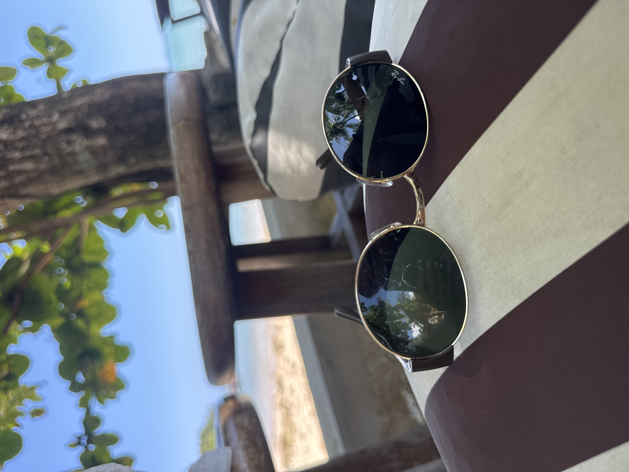 A pair of gold-rimmed, dark-lensed sunglasses rests on a brown and white striped cushion on a beach. The sunglasses reflect palm trees and the blue sky. In the background, a wooden table, a tree trunk, and a sandy beach with the ocean are visible under a bright blue sky with scattered clouds. The scene is set outdoors, likely in a tropical environment.