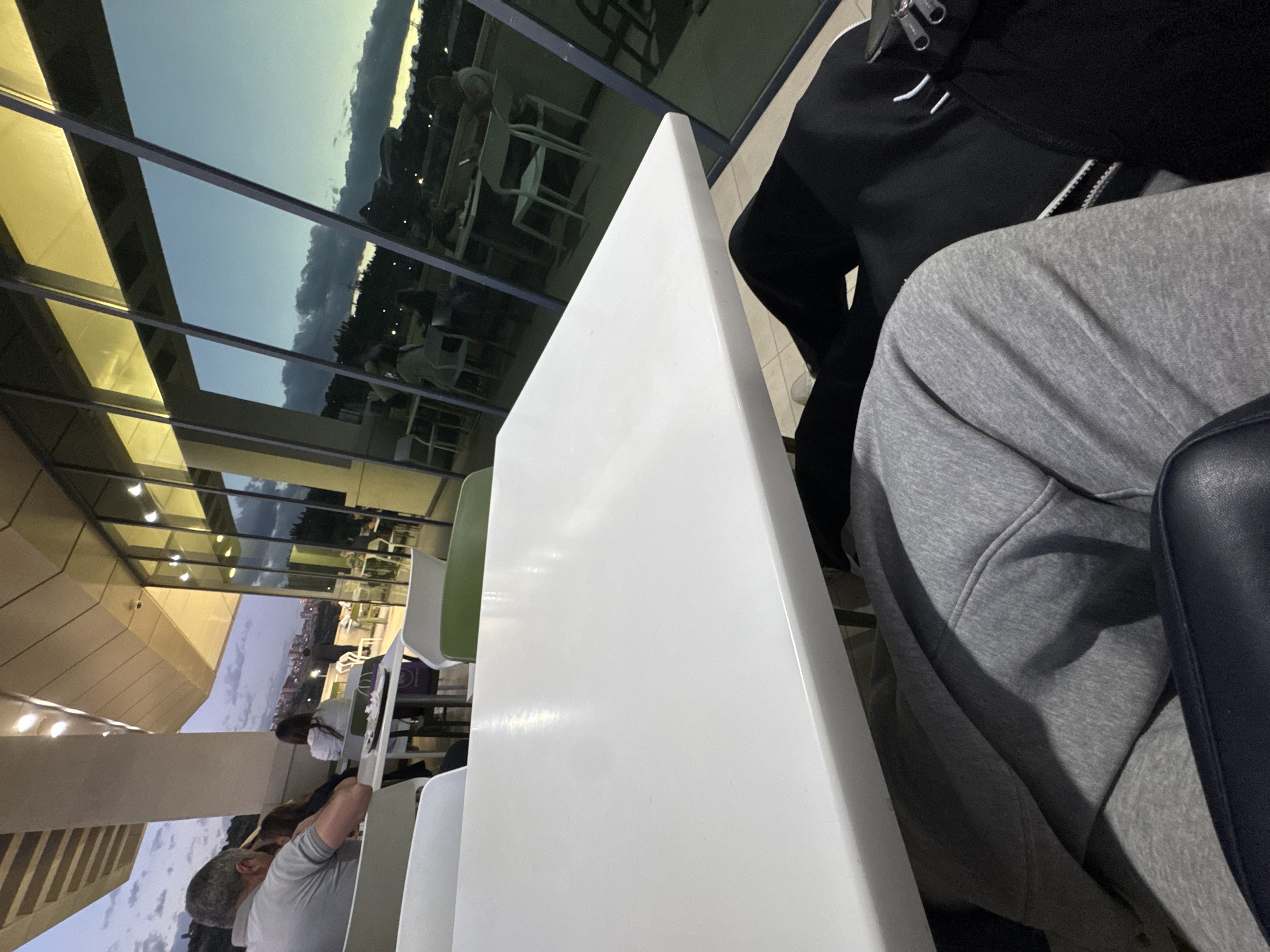 The image, taken with a portrait orientation and rotated, captures an indoor setting with a view of an outdoor cityscape at dusk in Yerevan, Armenia.
In the immediate foreground on the right, the lower torso and legs of a person wearing grey sweatpants are visible, alongside a black jacket or bag featuring white accents and zippers. Dominating the center foreground is a large, rectangular white table. To the left of the table, part of a white chair and a green chair are visible, suggesting a dining or lounge area.
The midground reveals other patrons seated at tables. On the left side, the back of a man with short grey hair is seen, leaning slightly forward. Further back, another person in a white top is seated at a table. The interior environment appears modern, with light-colored walls and ceilings, and contemporary furniture.
Through large, paneled glass windows in the background, a twilight sky with a mixture of clear patches and scattered clouds is visible. Below the horizon line, the subtle glow of city lights from Yerevan can be discerned, indicating an urban landscape. The overall lighting suggests it is early evening, with artificial lights illuminating the indoor space while natural light fades outside. The atmosphere seems relaxed and calm. No discernible text is visible on any objects or surfaces.
