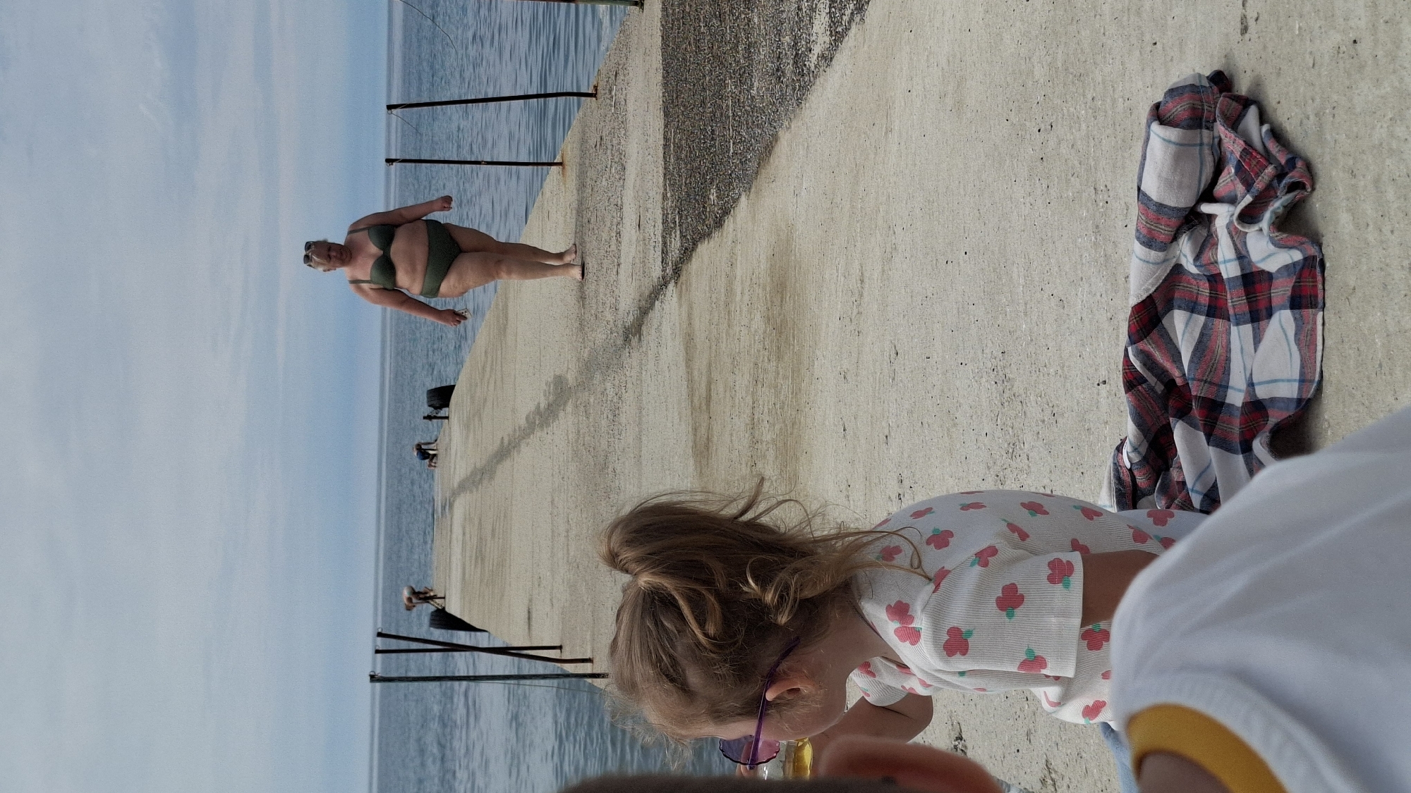 The image depicts a bright, sunny day at a pier or dock alongside a body of water, likely a lake or sea. The photograph is oriented vertically, showing the concrete pier extending into the distance on the right, with calm blue water and a clear sky to the left.
In the mid-ground, a woman with a fuller figure is standing barefoot on the concrete pier, wearing a dark green two-piece bikini and sunglasses. She has light-colored hair and is looking towards the camera with a neutral expression, her arms slightly bent.
In the immediate foreground, a young child with light brown hair tied in a ponytail is partially visible, facing away from the camera. The child is wearing a white long-sleeved top with small pink floral patterns and has sunglasses perched on their head or eyes. Their head is tilted downwards, suggesting they are looking at something on the ground or playing. To the right of the child, a red, white, and blue plaid shirt is casually draped on the concrete surface. Part of another person, possibly an adult, is visible in the very close foreground on the right, wearing a white shirt with a yellow trim, out of focus.
The water appears calm, reflecting the light sky, and faint figures of other people can be seen further down the pier in the distance, alongside dark poles or pilings extending from the water. The weather is clear and bright, indicating daytime. There is no visible text in the image.