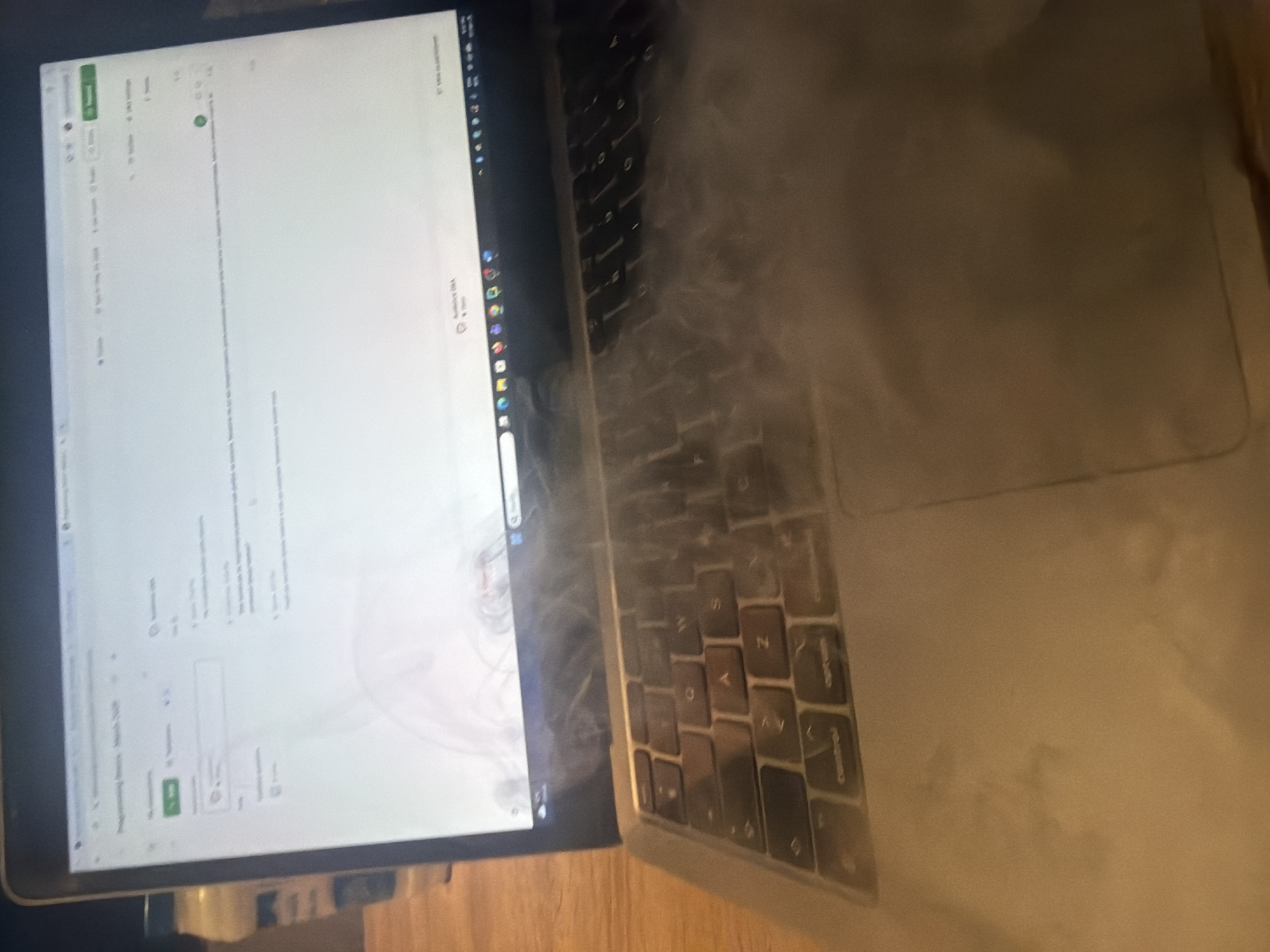 A laptop, partially obscured by smoke, is positioned on a wooden surface. The laptop screen displays what appears to be a word processing or document creation interface. The laptop keyboard and surrounding area are heavily affected by the smoke. The environment suggests an indoor setting. No information about Plovdiv, Bulgaria is available. No people or interactions are visible in the image.