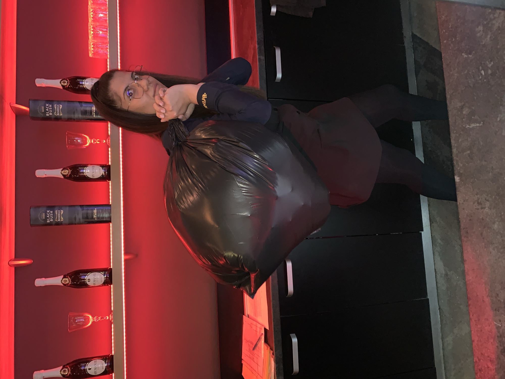 The image shows a young woman with dark hair, wearing glasses, and holding a large black trash bag over her shoulder. She is wearing a dark blue long-sleeved shirt with an orange logo on the sleeve, and a dark red skirt or apron. The background features a shelf with bottles of alcohol and glassware, lit by red lighting. Below the shelf are black cabinets with silver handles. On the counter to the left, there are papers and a pen. The scene appears to be indoors, possibly a bar or a home bar. The location is not identifiable from the image alone, and there is no information about Burgas, Bulgaria.