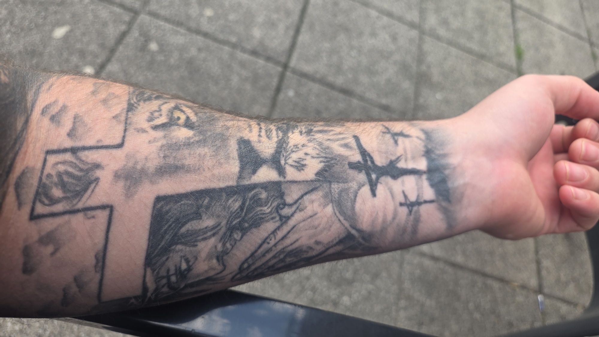 A close-up shot reveals a person's forearm adorned with a black ink tattoo. The tattoo features a cross, the eye of a lion, an eagle and barbed wire. The background shows grey paving stones. The location is unknown.