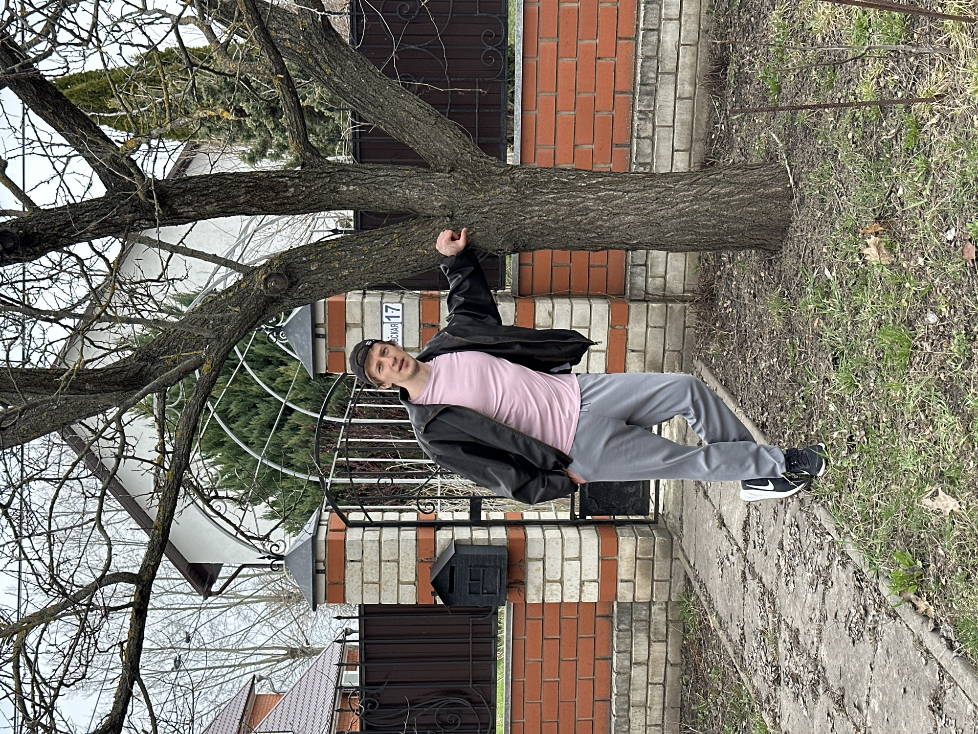 A man stands next to a tree in the city of Kursk, Russia. He is wearing a black jacket, a pink t-shirt, grey sweatpants, and black and white sneakers. He is also wearing a black baseball cap. He has his left hand on the tree. The tree trunk is horizontal and runs along a brick and metal fence. The ground is covered in grass and leaves.