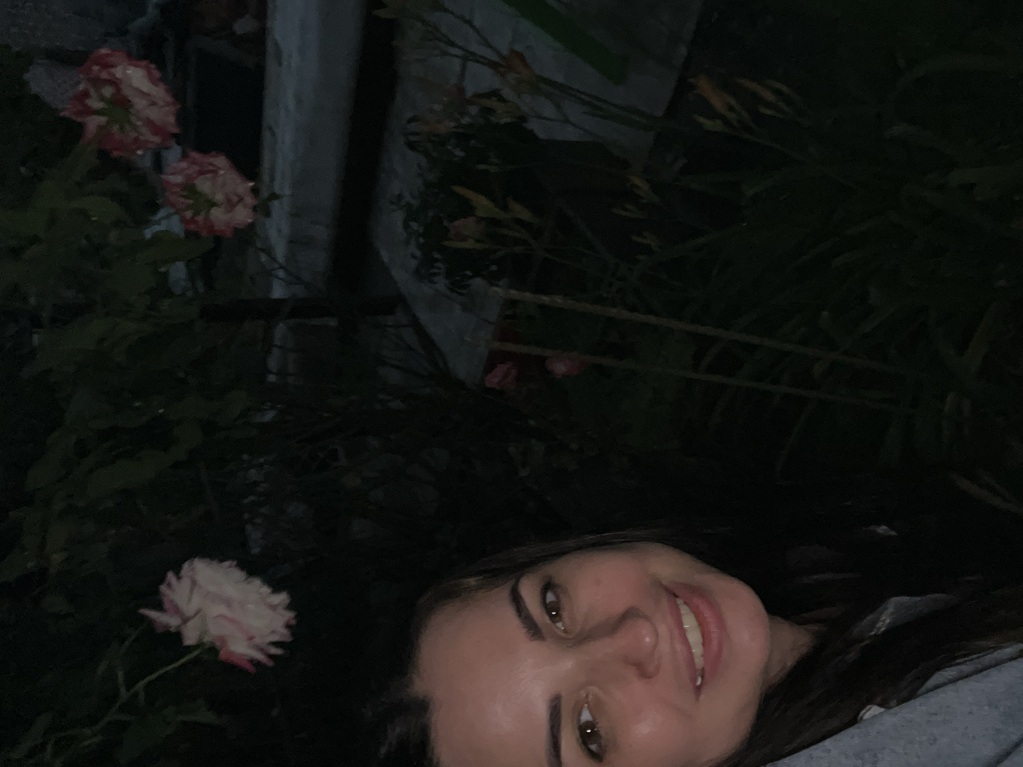 An outdoor, low-light, close-up selfie shows a woman's face from the chest up, smiling. She has brown hair and brown eyes, and is wearing a light blue collared shirt. Behind her, roses with white and pink petals are visible, as are other flowers and dark green foliage. The background is dark, suggesting it is nighttime. The location is listed as Unknown, Unknown.