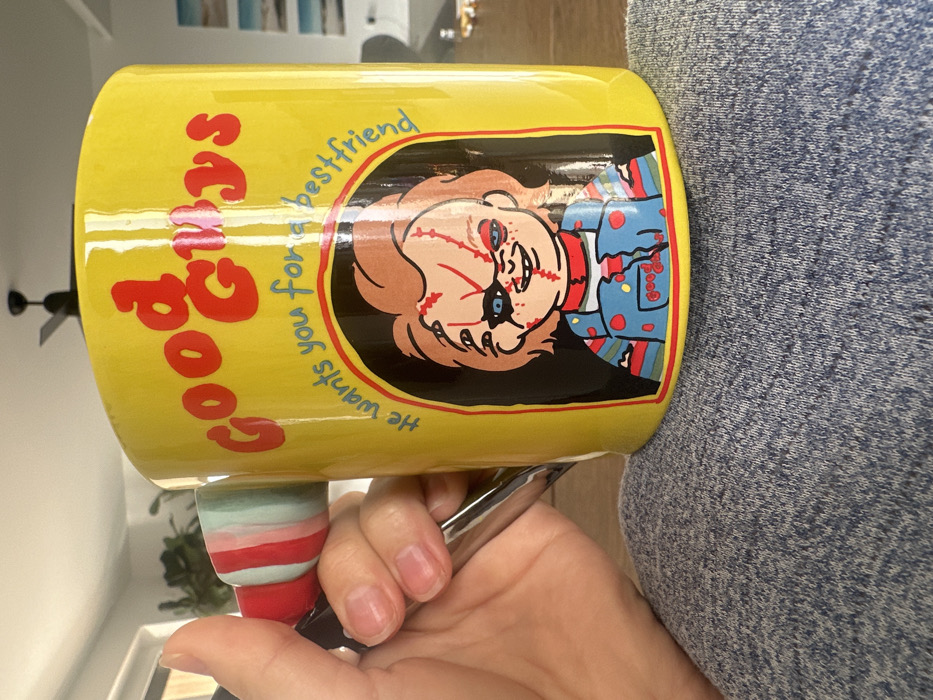 The image displays a close-up, angled view of a person's right hand holding a yellow ceramic mug, observed within an indoor setting in Ericeira, Portugal. The mug, prominent in the foreground, features a cartoon rendition of the "Good Guys" Chucky doll. Red text at the top of the mug reads "Good Guys," with smaller, curved text below stating, "He wants you for a bestfriend." The Chucky illustration depicts a doll with red hair, facial scars, one blue eye, and a dark patch over the other, dressed in torn blue overalls over a striped shirt. The mug's handle exhibits a vertical striped pattern in red, light blue, and white, with a dark, possibly metallic, base.
The person's right hand grasps the handle, with visible fingernails that appear natural and unpainted. A subtle metallic ring is present on the ring finger. Below the mug, the person's lap is visible, covered in greyish-blue heathered fabric, indicative of casual attire. The background is softly blurred, revealing a dark four-bladed ceiling fan at the top, and indistinct rectangular shapes on a distant wall, possibly artwork or windows, with muted colors suggesting landscapes. A dark, possibly wooden, surface is partially visible in the lower right background.
