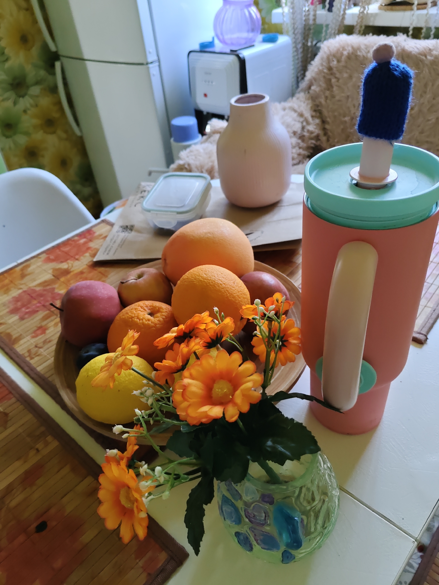 This image captures a still life on a table, likely in a domestic setting in Rostov-on-Don, Russia. In the foreground, a round, light-brown wooden bowl contains an assortment of fruits, including two oranges, one red apple, a yellow lemon, and a dark plum. Interspersed with the fruit are artificial orange flowers with green stems and some small white buds. This arrangement sits on a placemat with a floral pattern. To the right of the fruit bowl is a tall, pink tumbler with a light green lid and a pale beige handle. Atop the tumbler, a small, dark blue knitted cover with a light brown pom-pom sits on a white cylindrical object. In the background, a light pink vase with a rounded base and a narrow neck is visible, as is a white refrigerator with a floral curtain hanging next to it. Various kitchen appliances, including a white unit with a blue knob, are also present in the background. The lighting suggests an indoor environment, possibly daytime.