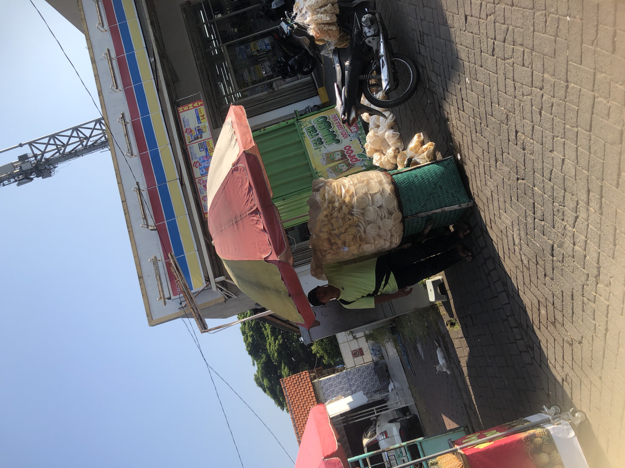 A man stands next to a stall selling snacks in front of a convenience store. The stall is covered by a red and yellow umbrella and is piled high with bags of chips. Behind the stall, a motorcycle is parked. In the background, a white car is parked next to a building with a red tiled roof. The scene is set on a sunny day with a clear blue sky. The convenience store appears to be a branch of Indomaret, a popular Indonesian convenience store chain, based on the signage. The overall setting suggests a street-side vendor operating in a commercial area of Kalisampurno, Indonesia.