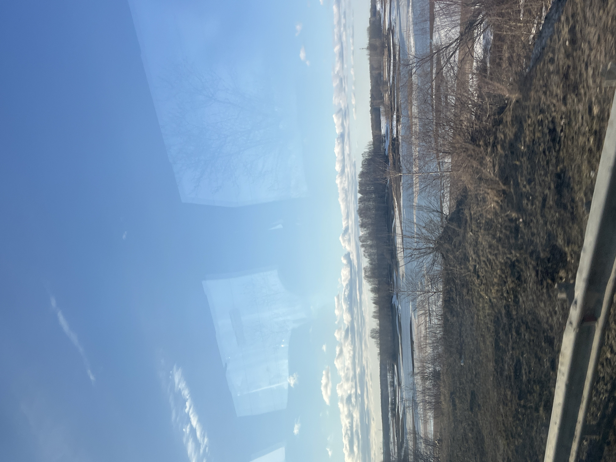 The photograph depicts a view from a vehicle's window. The left side of the image features a clear blue sky with some wispy clouds and a reflection of trees or structures. The right side of the image shows a landscape featuring a body of water partially covered with ice or snow, bordered by trees and vegetation. A guardrail runs along the bottom right corner. The overall impression is a wintery or early spring scene. The city of Berezniki, Russia, is not explicitly identifiable in the photograph. The landscape could be located in any geographic region with similar characteristics. Without more context, pinpointing the exact location is not possible.