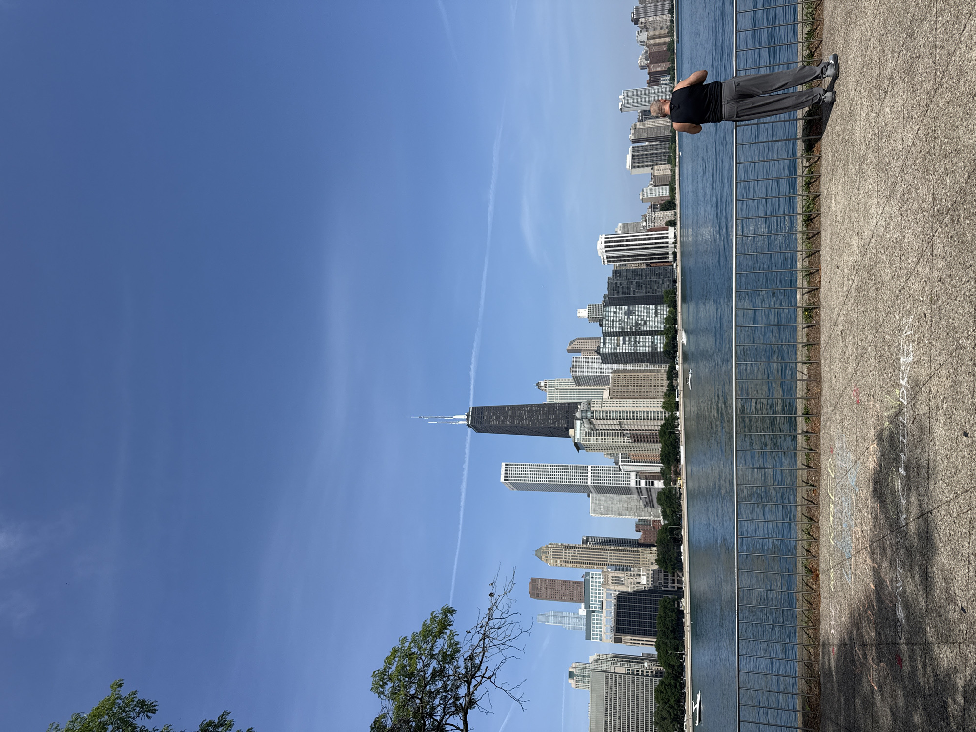 This is a vertically oriented, eye-level photograph of the Chicago skyline. The image is dominated by a clear, bright blue sky. In the foreground, a paved walkway is visible, with a metal railing separating it from a body of water. A man, seen from behind, is standing on the walkway, facing the water and the skyline. He is wearing a black tank top and grey pants. His arms are bent as if he is performing a push-up or resting in a plank position. The Chicago skyline is in the background, featuring numerous tall buildings, including the distinctive John Hancock Center. Trees are visible along the waterfront, and a few boats are on the water. The lighting suggests it is a sunny day, likely late morning or early afternoon, given the bright sunlight and shadows. The scene conveys a sense of urban recreation and appreciation for the city's architecture and waterfront. There is no visible text in the image.