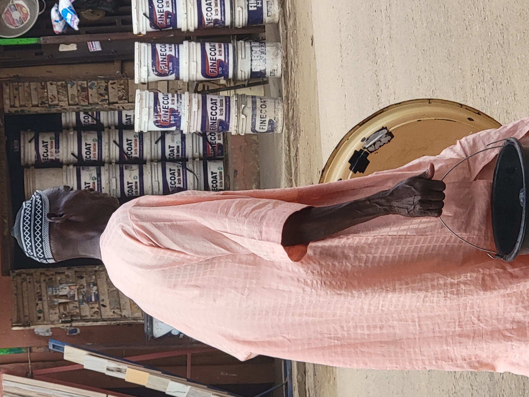 This image, captured in Maiburiji, Nigeria, portrays an elderly individual in what appears to be a paint or hardware store. The person, viewed from the side, wears a light peach-colored, long-sleeved garment and a distinct black and white patterned cap. Their right arm, with visible wrinkles, holds a small black bucket by its wire handle.
The background is dominated by shelves stacked high with numerous white cans of paint. "FINECOAT" and "ECOAT" are clearly legible brand names on these cans, with "Acrylic Emulsion" also visible on some labels, indicating the type of paint. Below these main stacks, other shelves display small, rectangular samples, possibly paint swatches or tile samples. To the right, a large, yellow, circular metal object, potentially a basin or part of a wheel, is partially visible against a light-colored wall.
The scene is brightly lit by natural daylight, suggesting it is daytime with clear weather. The individual's posture is calm, looking slightly upwards or into the distance. The setting and visible products suggest a local commercial establishment where building materials or home improvement items are sold.