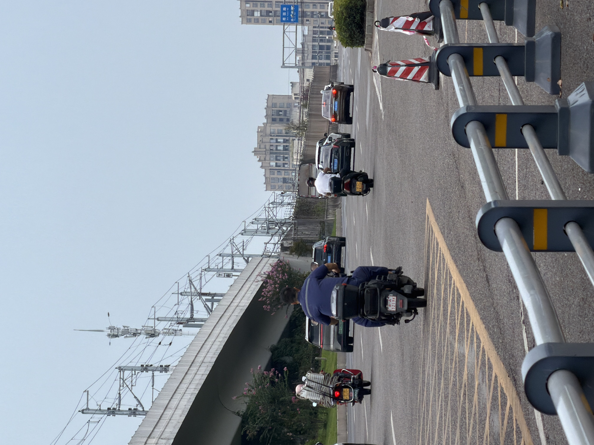 The image depicts a street scene in the city of Jiangjin, China, characterized by a multi-lane road under an elevated structure with power lines. Several vehicles are visible, including a red scooter carrying a load of what appears to be coiled wire and other items, a black scooter with a rider in a blue uniform, and various cars. The road has yellow lane markings. In the background, there are buildings and more power lines. Traffic cones and a metal barrier are present along the side of the road. The sky is overcast.