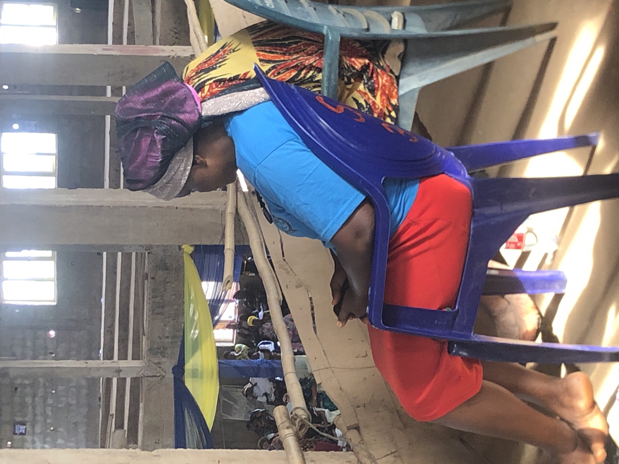 An adult in Nteje, Nigeria, wearing a purple head covering, a light blue t-shirt, and red shorts is partially seated in a blue plastic chair. The scene appears to be an indoor gathering of people seated on mats. In the background, structural elements of a building are visible, along with yellow and blue drapery. A partially visible window is on the upper-left. Other chairs are present in the foreground. The image is rotated 90 degrees clockwise.