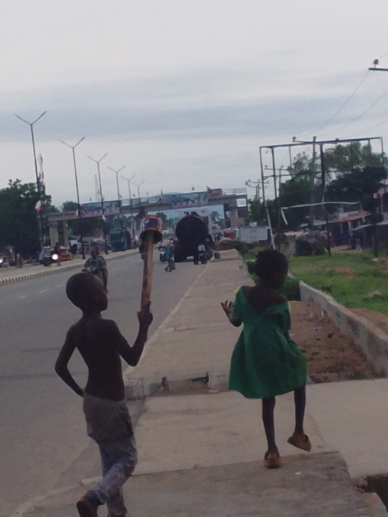 The image depicts a street scene in Jalingo, Nigeria, on an overcast day. In the foreground, two young children are visible. The child on the left, a boy, is facing away from the camera, holding a long object with an orange and black top aloft in his right hand. He is wearing light-colored, patterned trousers. The child on the right, a girl, is also facing away from the camera, wearing a green dress. She is positioned slightly further down the sidewalk.
Behind the children, a paved road with a white median line is visible, with vehicles and other pedestrians in the middle ground. Several tall, modern streetlights are spaced along the roadside. A large, dark structure, possibly a truck or bus, is also present on the road. Further in the background, a bridge-like structure with decorative elements spans the road, and beyond that, trees and buildings can be seen. The environment appears to be an urban setting with infrastructure typical of a Nigerian city. The overall atmosphere is subdued, likely due to the cloudy weather.