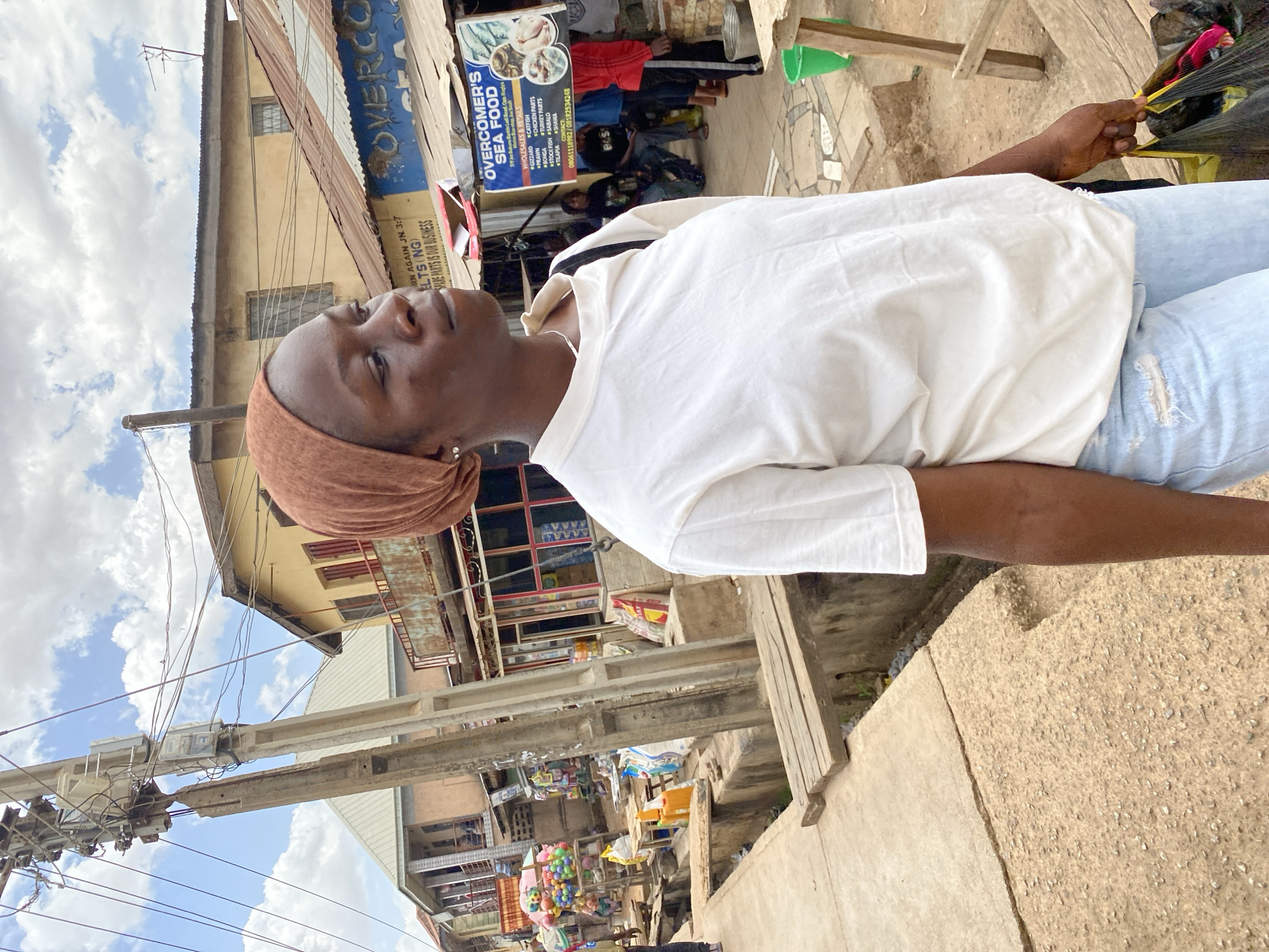 The image depicts a woman standing on a sidewalk in a busy urban environment, likely Bukuru, Nigeria, based on signage and visual cues. She is wearing a white t-shirt, light-colored denim jeans with a rip, and a brown head covering. She is holding a black trash bag filled with items. The background shows storefronts and buildings, including one with a sign that reads "OVERCOMER'S SEA FOOD" listing various seafood items. There is a power pole with electrical equipment visible against a cloudy sky. People are visible in the background, further suggesting a bustling commercial area. The ground is concrete with varying textures, indicating repairs or different construction phases.