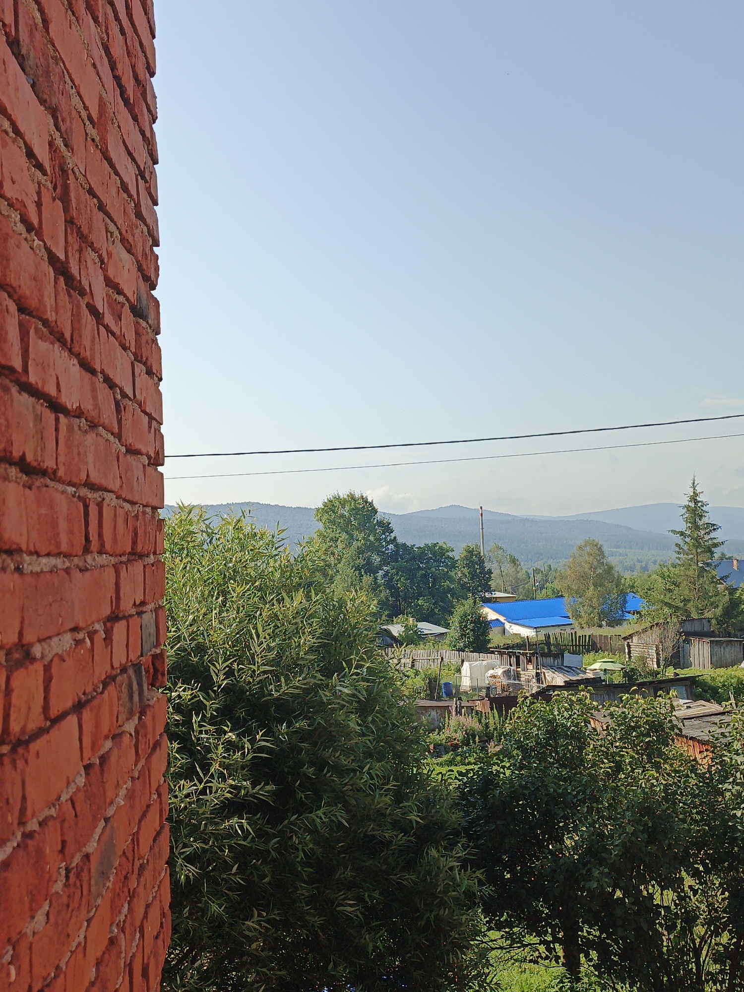 This is a medium shot taken from an elevated perspective looking east over the village of Medvedyovka, Russia. In the foreground on the left, a red brick wall is visible, taking up approximately one-third of the frame. Beyond the wall, a lush green tree with thin leaves dominates the mid-ground. Further in the distance, a cluster of rural houses with various roof types, including a prominent bright blue roof, are nestled among trees. A tall, slender smokestack with a red and white top is visible behind the houses. The background is comprised of rolling hills covered in dark green trees, fading into a hazy blue sky. A thin black power line stretches horizontally across the upper middle portion of the image. The scene appears to be a sunny day with clear, light blue skies.