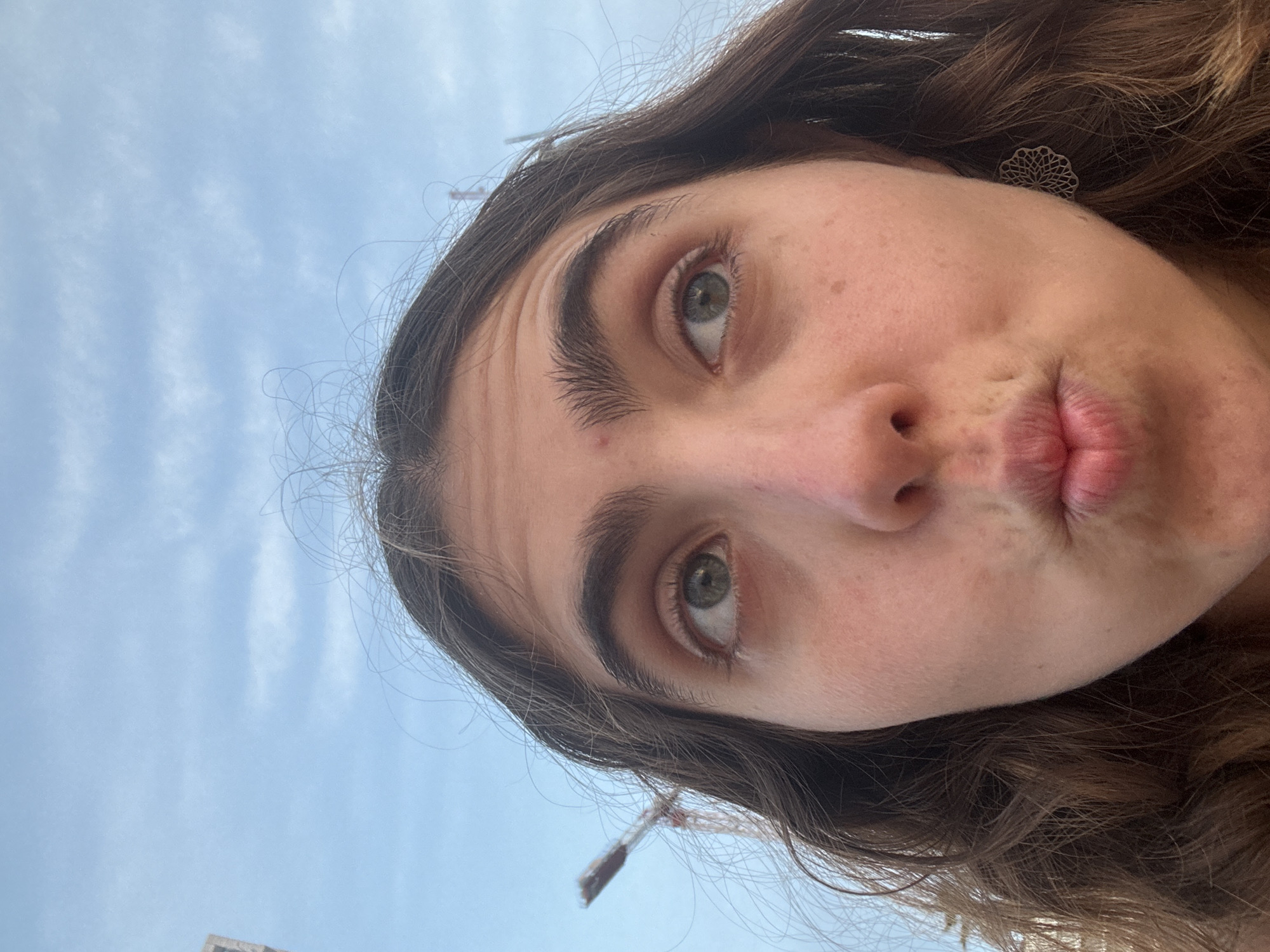 A close-up selfie shows a woman with light skin, brown hair, and green eyes, looking up at the camera with pursed lips. She is wearing a silver, flower-shaped earring. Behind her is a light blue sky with wispy clouds. In the lower left corner, there's a glimpse of a building and a construction crane. Based on the architectural style, it is possibly within the city of Tel-Aviv, Israel.