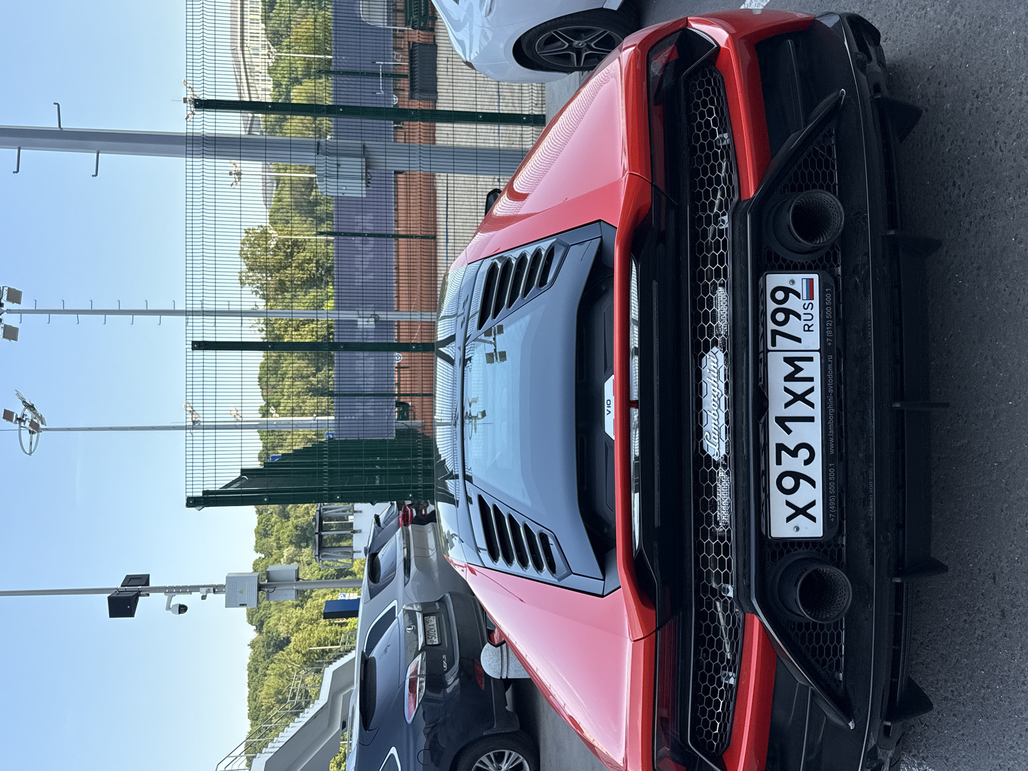 This media file shows a close-up of the rear of a red Lamborghini parked outdoors. The car is the primary subject, with its distinctive taillights, exhaust pipes, and rear diffuser clearly visible. The license plate, which reads "X931XM 799," is also prominent. The car appears to be in good condition, with a shiny red paint job.
The background suggests a sunny day, with a clear blue sky visible above. A green metal fence runs along the left side of the frame, with trees and buildings visible behind it. The ground is paved, indicating a road or parking lot. Other cars are partially visible in the background, including a white car and a darker colored car.
The overall scene is one of a parked supercar, likely in a public area or showroom. There are no people visible in the frame. The text "Lamborghini" is visible on the car, along with website and phone number details that suggest it may be a dealership or service center. The time of day appears to be daytime due to the lighting. The location is suggested to be Moscow, Russia by the Cyrillic text on the license plate.