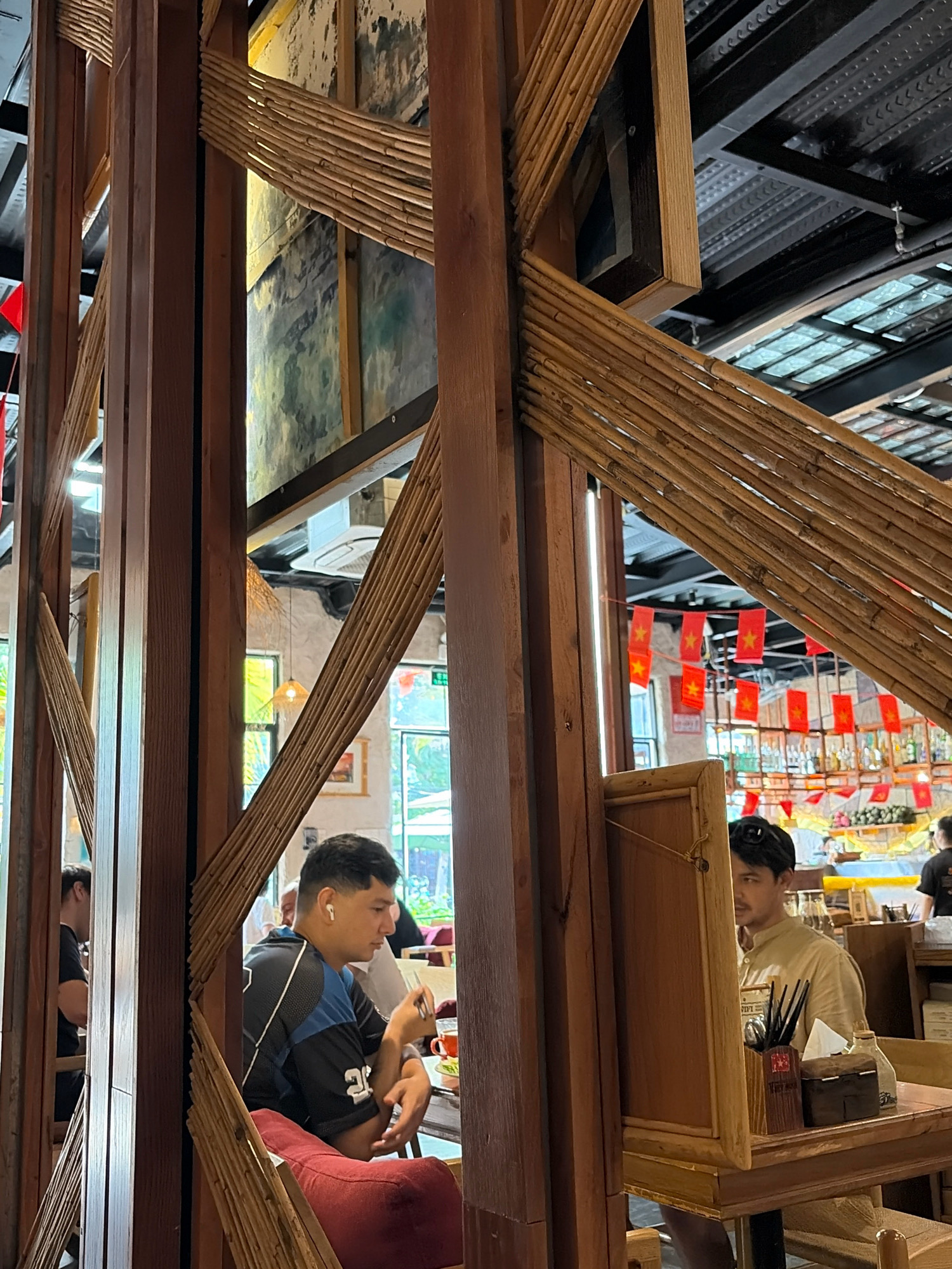 The image depicts the interior of a restaurant with a rustic and natural design, likely located in Đà Nẵng, Vietnam, as suggested by the Vietnamese flag decorations. The structure of the restaurant is characterized by prominent wooden beams and diagonal bamboo elements that create an open, somewhat partitioned space. In the foreground, two men are seated at tables. The man on the left, wearing a black and blue t-shirt and white earbuds, is leaning forward, appearing to be engaged in a meal or conversation. The man on the right, in a light-colored shirt, is looking towards the left, with a wooden partition obscuring part of his face. The background reveals a bar area with shelves stocked with bottles and more red flag decorations with yellow stars, common in Vietnam. The lighting appears to be a mix of natural light filtering through windows and artificial overhead lighting, contributing to a warm ambiance.
