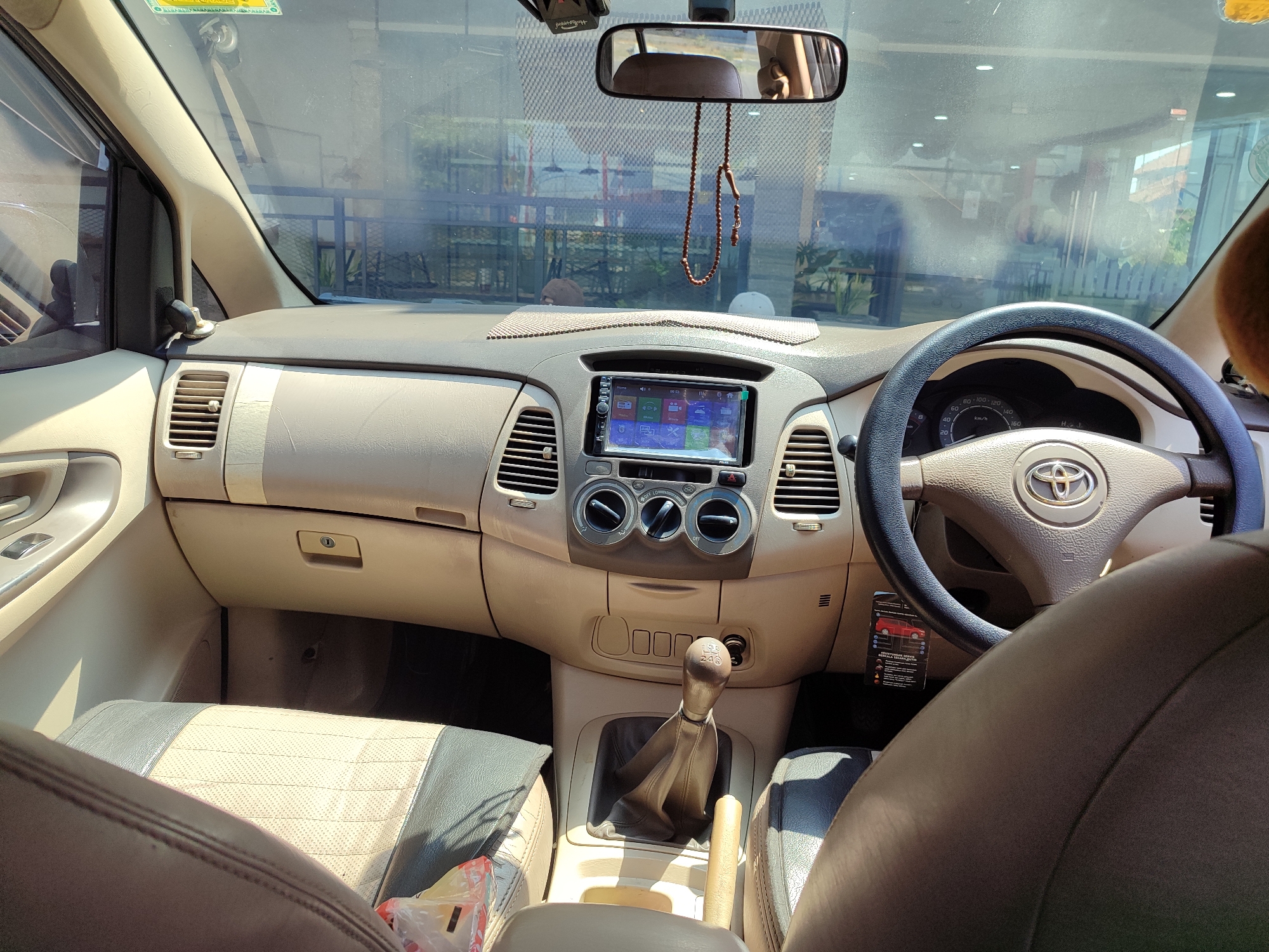 The image shows the interior of a car, viewed from the passenger seat. The dashboard is tan and features a touchscreen infotainment system, air vents, and climate control knobs. A steering wheel with a Toyota logo is visible on the right. The gear shifter is in the center console, and a handbrake is to its right. The seats are covered in a tan and grey fabric. A string of wooden beads hangs from the rearview mirror. The background visible through the windshield suggests an outdoor setting with some fencing and greenery, possibly a parking lot or a street in Tegal, Indonesia. The sun is shining, casting light and shadows within the car.