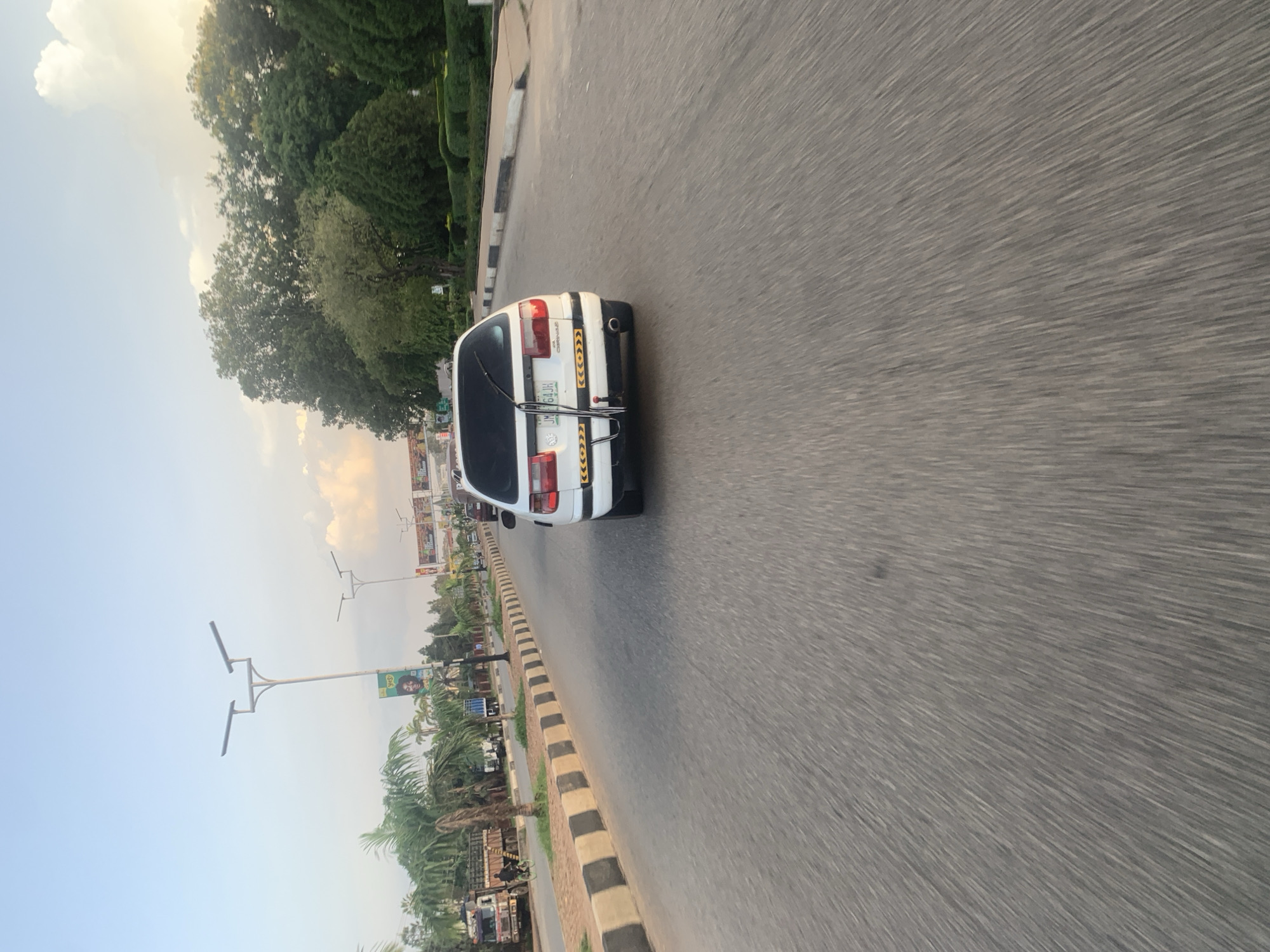 The image depicts a street scene in Jos, Nigeria, at what appears to be late afternoon or early evening due to the soft lighting and long shadows. A white sedan is traveling forward on a paved road, captured from a perspective behind and slightly above the vehicle. The car has a license plate visible, and a black object, possibly a bicycle rack, is attached to its rear. The road is flanked by a median strip with black and beige curbing, and beyond that, a sidewalk area with palm trees and manicured bushes. In the background, there are more trees and some buildings or structures, including what appear to be streetlights and billboards. The sky is a pale blue with some clouds, suggesting fair weather. The motion blur effect on the road indicates that the photo was taken from a moving vehicle. Further down the road, other vehicles and possibly pedestrians can be observed, but they are not clearly discernible due to the distance and motion blur. The overall impression is a snapshot of everyday life on a road in Jos.