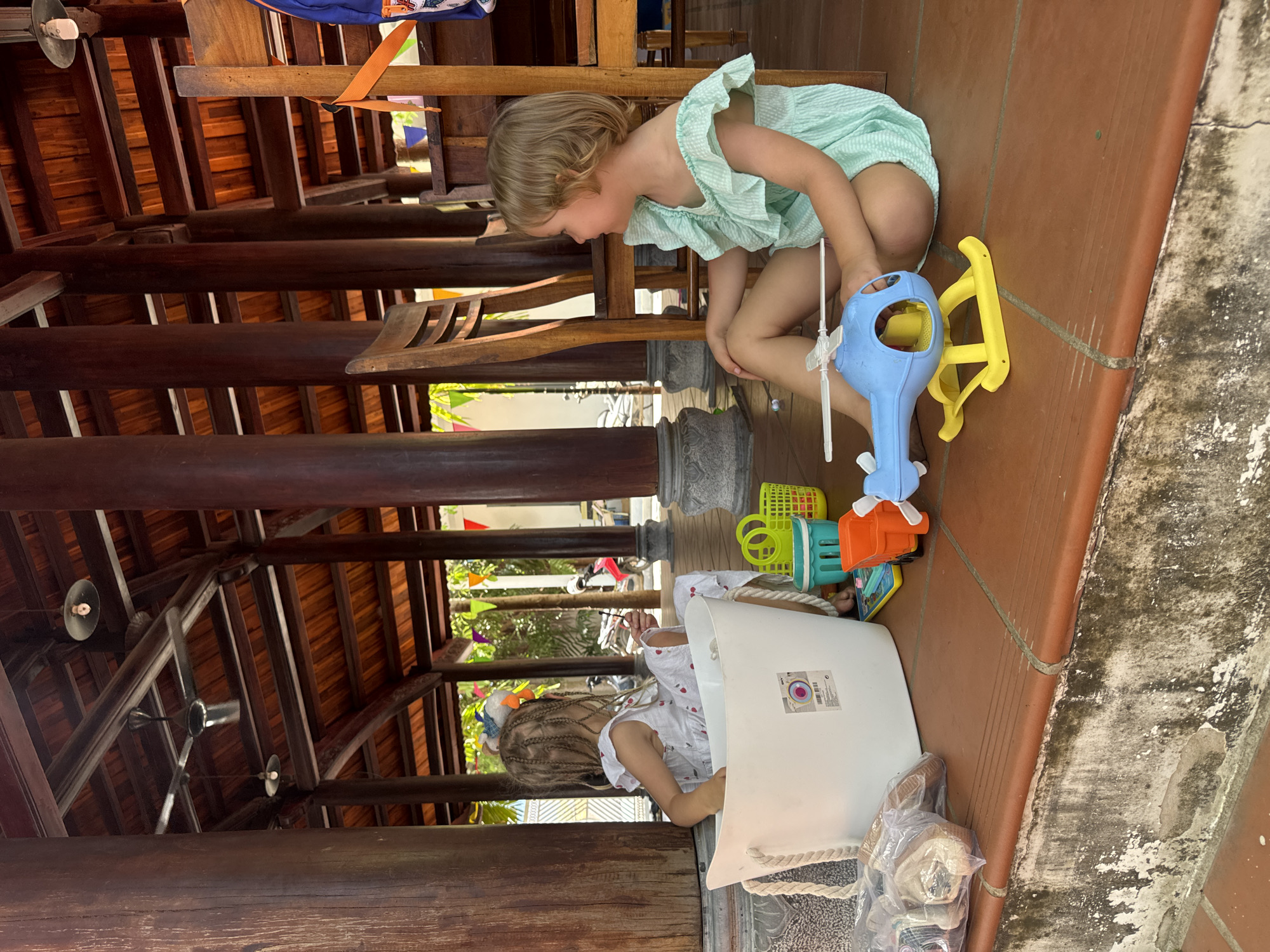 Two young girls are playing with toys indoors. One girl, with short blonde hair, is wearing a green outfit and is crouched down playing with a blue and yellow toy helicopter. The other girl, who has her hair in braids, is wearing a white dress and is looking into a large white bucket filled with toys. The scene is inside a building with dark wooden pillars and beams. The floor is tiled in a reddish-brown color. There is a view to the outside through the pillars, where some greenery and other buildings can be seen. The location is Mũi Né, Vietnam, evidenced by the architectural style of the building.