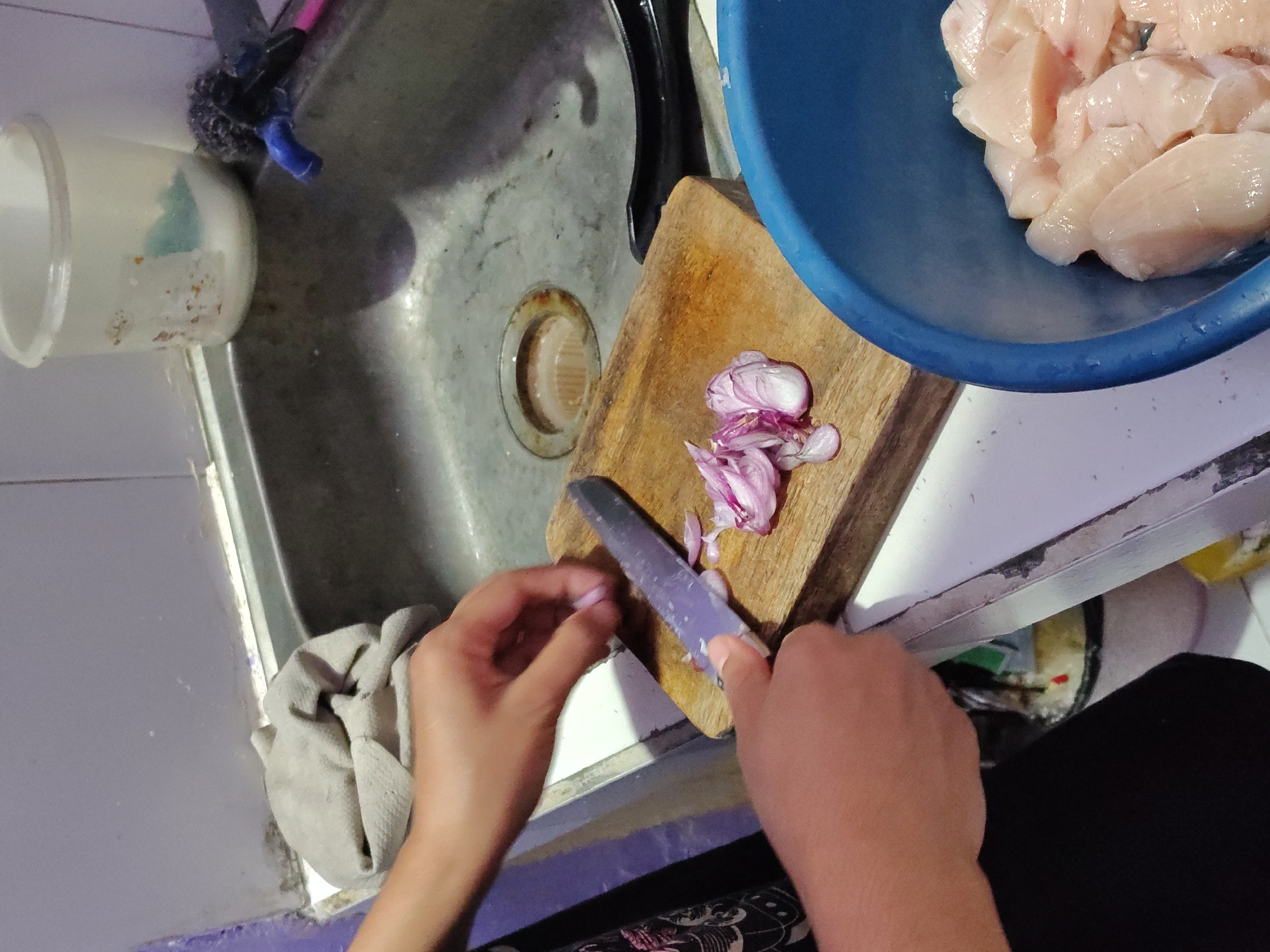 A person is chopping red onions on a wooden cutting board next to a kitchen sink. The sink is dirty with rust and debris. A blue bowl filled with raw chicken pieces is to the right of the cutting board. In the background, a dirty white plastic container and a faucet are visible. A gray cloth is draped over the edge of the sink. The scene appears to be in a home kitchen in Surabaya, Indonesia.