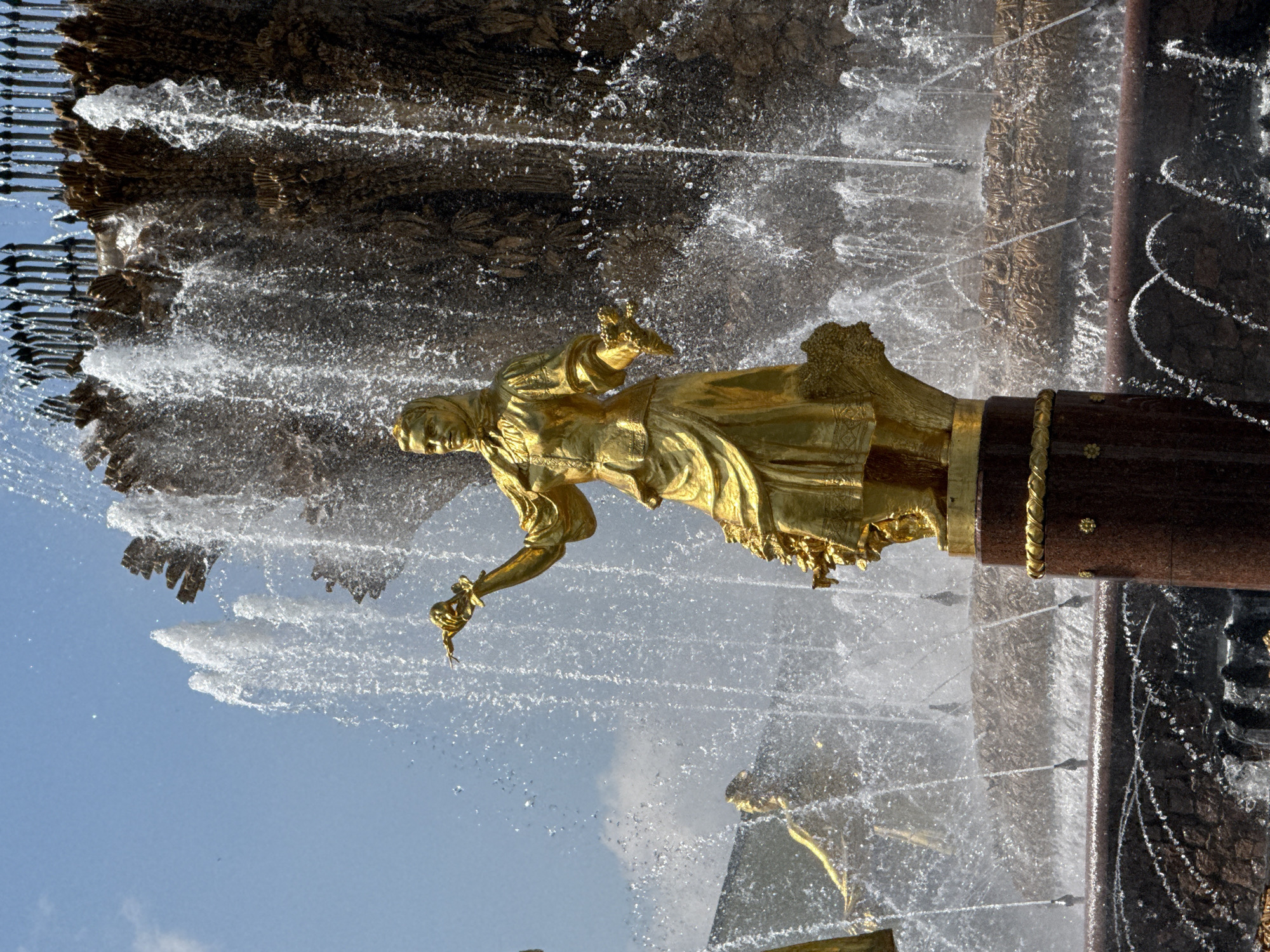 This image shows a close-up view of a golden sculpture, possibly made of bronze, located in Moscow, Russia. The sculpture depicts a human figure, clad in a long dress or robe, standing atop a pedestal or column. The fountain is in full operation, with water cascading and spraying around the sculpture. The background consists of a combination of the sky. The location could be the "Friendship of Nations" fountain at the Exhibition of Achievements of National Economy (VDNKh) in Moscow. The angle of the shot is from a low perspective, looking upwards at the statue.