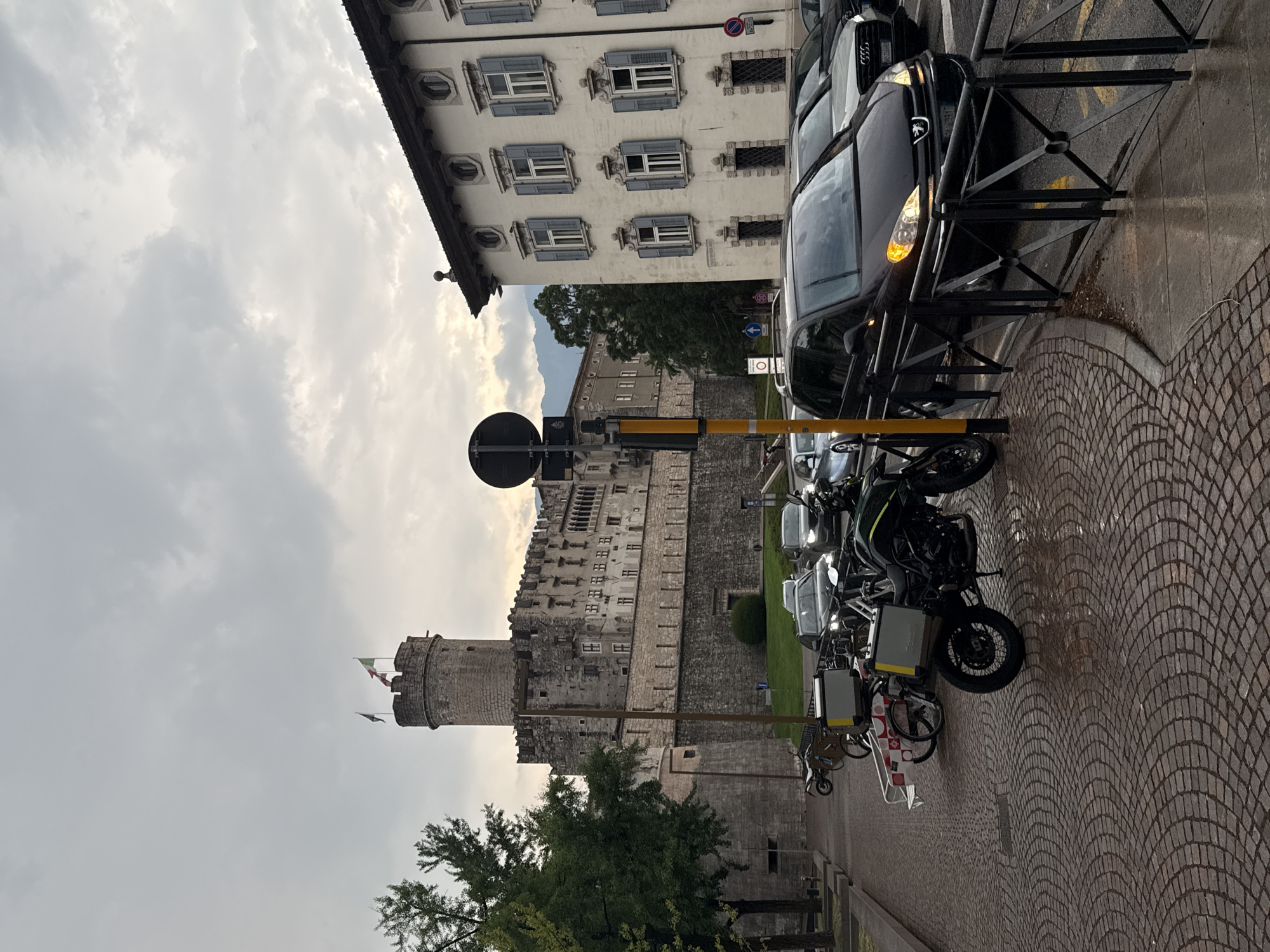 The image depicts a view of the Buonconsiglio Castle in Trento, Italy. The castle, a large stone structure with a prominent tower, stands in the background. In the foreground, a street scene unfolds with several parked motorcycles and a trailer, as well as cars on the wet road. The architecture of a building to the right of the castle is also visible, characterized by its light-colored facade and shuttered windows. The sky is overcast with clouds.
