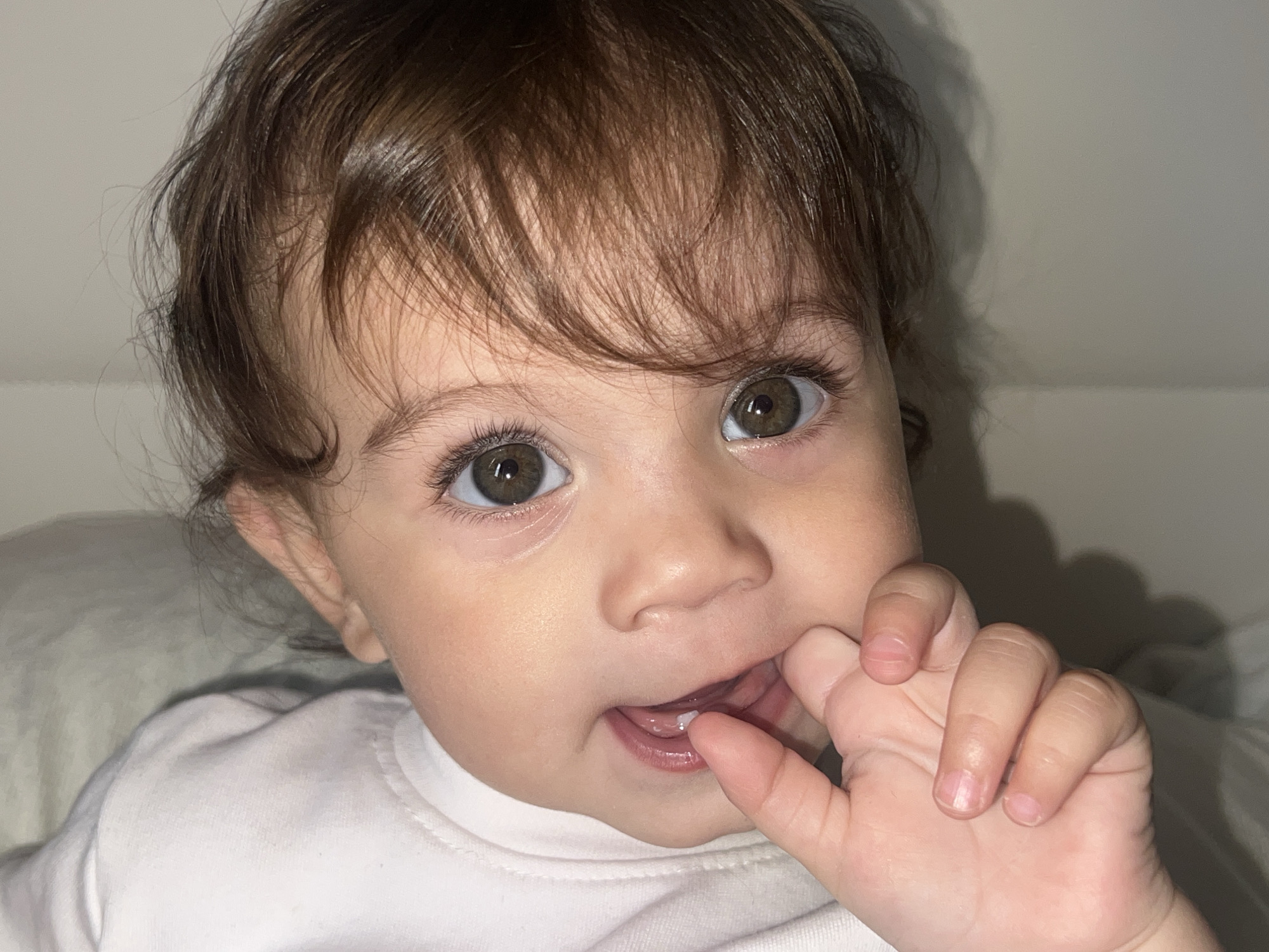 A close-up shot depicts a young child with brown hair and hazel eyes. The child's right hand is held up to their mouth, with a finger partially inside. The child is wearing a white shirt and appears to be indoors. The background is a neutral color. There's no specific information to indicate the location is Elena, Bulgaria, or to connect the image to that city.