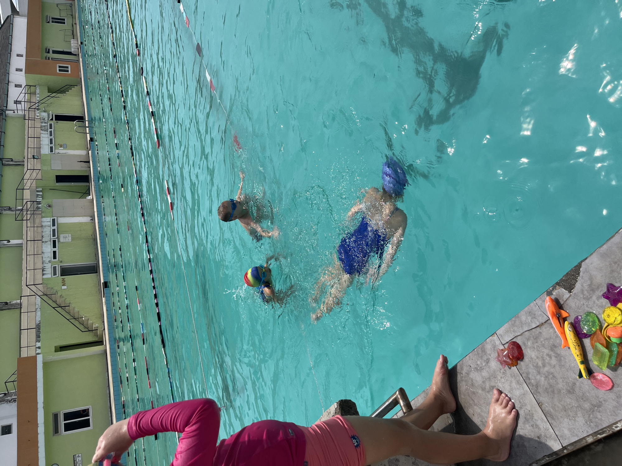 The image shows an outdoor swimming pool with three children swimming in it. One child is wearing a blue swimsuit and swim cap, and another child is wearing goggles. The third child is wearing a colorful swim cap and goggles. The pool has lane markers and is surrounded by a building with green walls. A person is sitting on the edge of the pool with their feet dangling in the water. There are several colorful plastic toys on the pool deck. The location appears to be Nha Trang Ward, Vietnam, based on the "NAM" sign on the building.